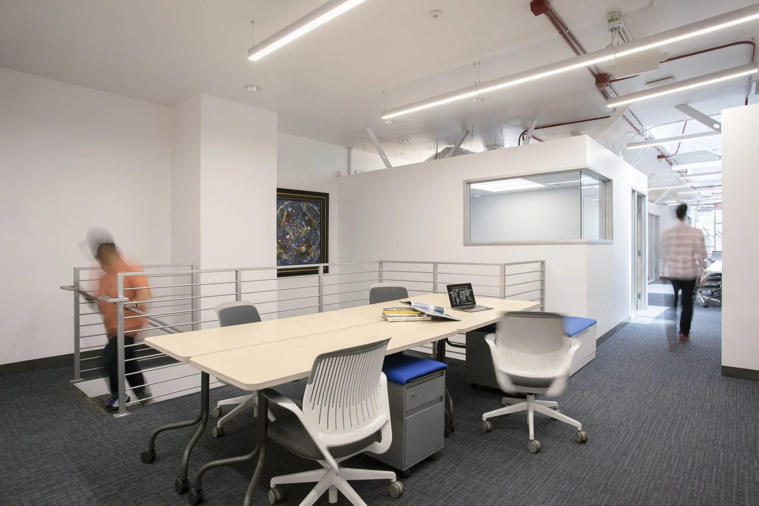 Empty office space with a large table and chairs, one bicycle behind a railing, a framed abstract painting on the wall, and two people walking in the background.