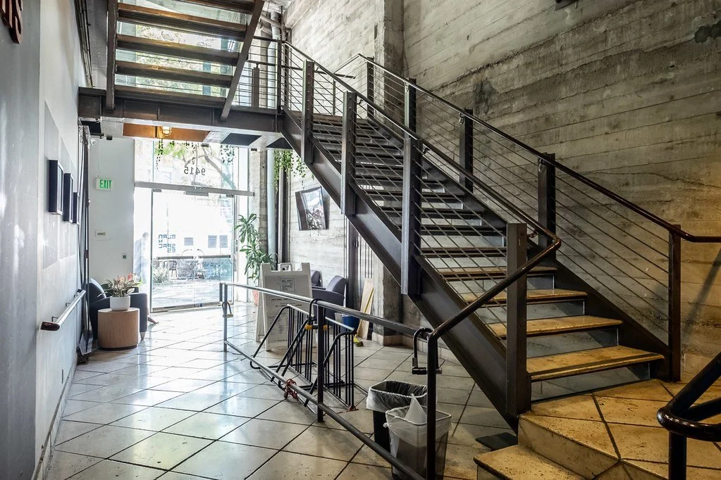 Interior of a modern building with concrete walls, a metal staircase with wooden steps, tiled floor, and natural light coming through the glass front door.