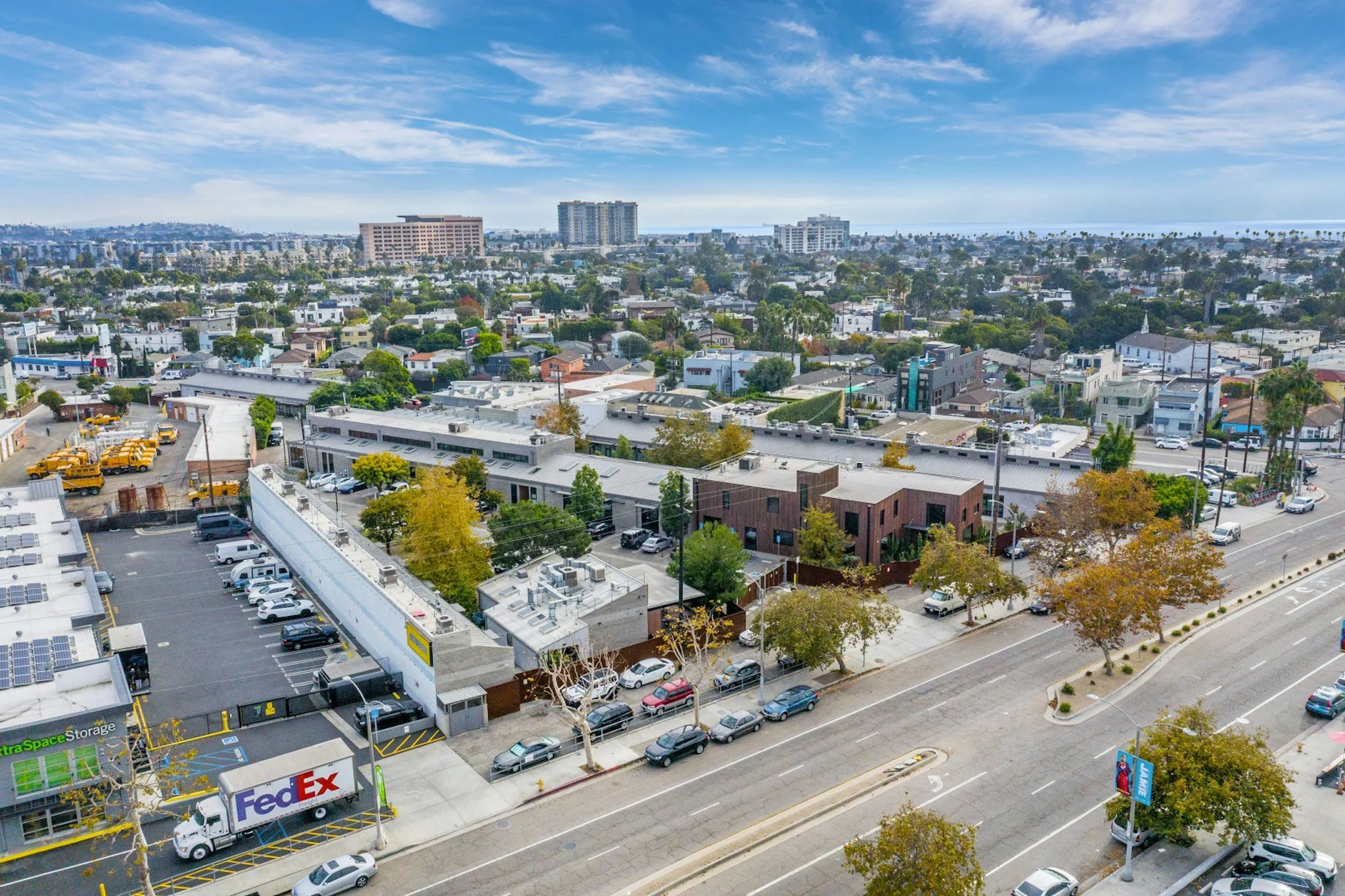 Aerial view of an urban area with a mix of commercial and residential buildings, parking lots, and trees under a partly cloudy sky, with a distant view of the ocean.