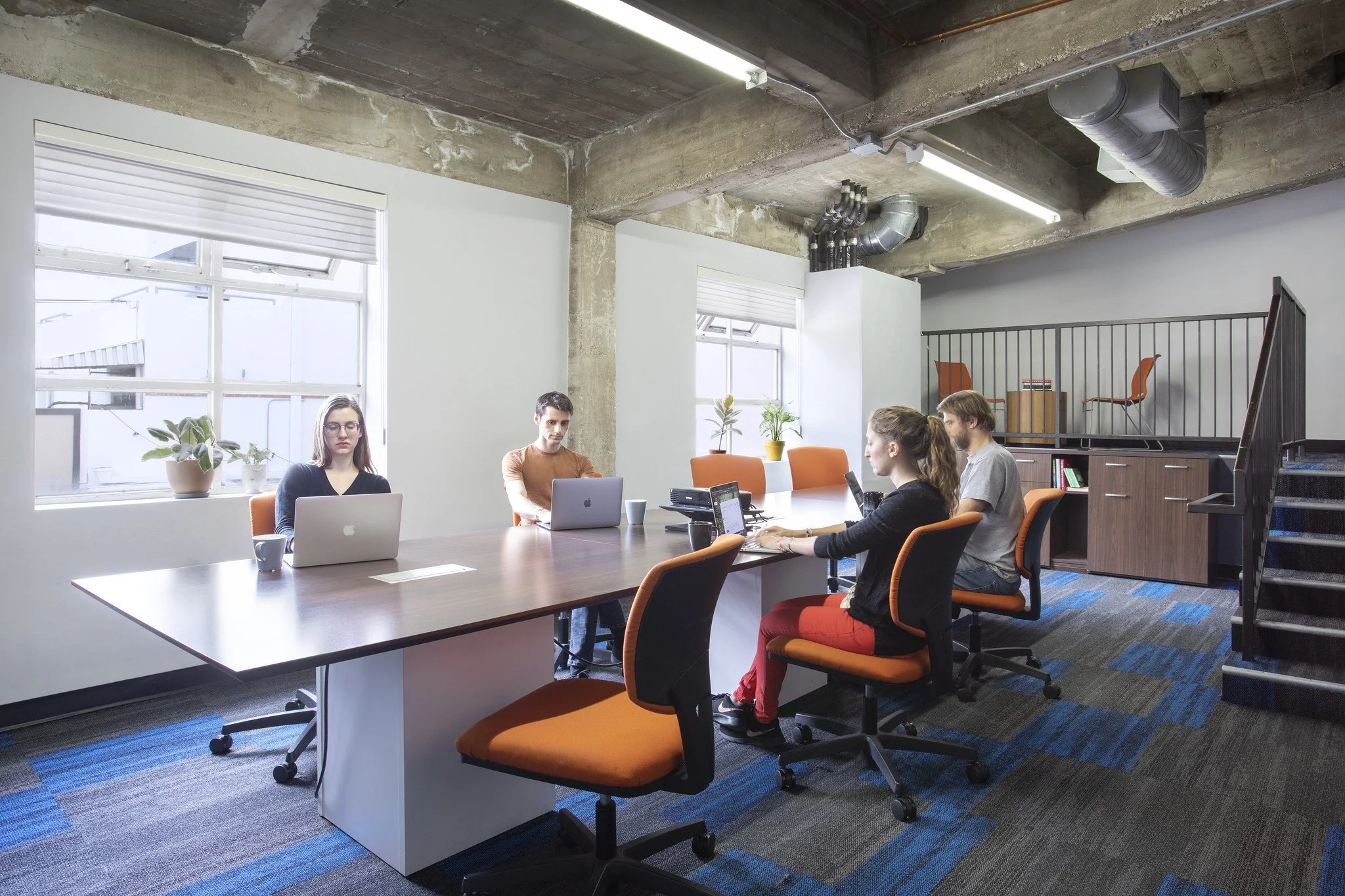 A modern office space with five people working at a large table, laptops open, and windows allowing natural light. The room has an industrial style with exposed concrete and ductwork, and orange chairs with black frames.
