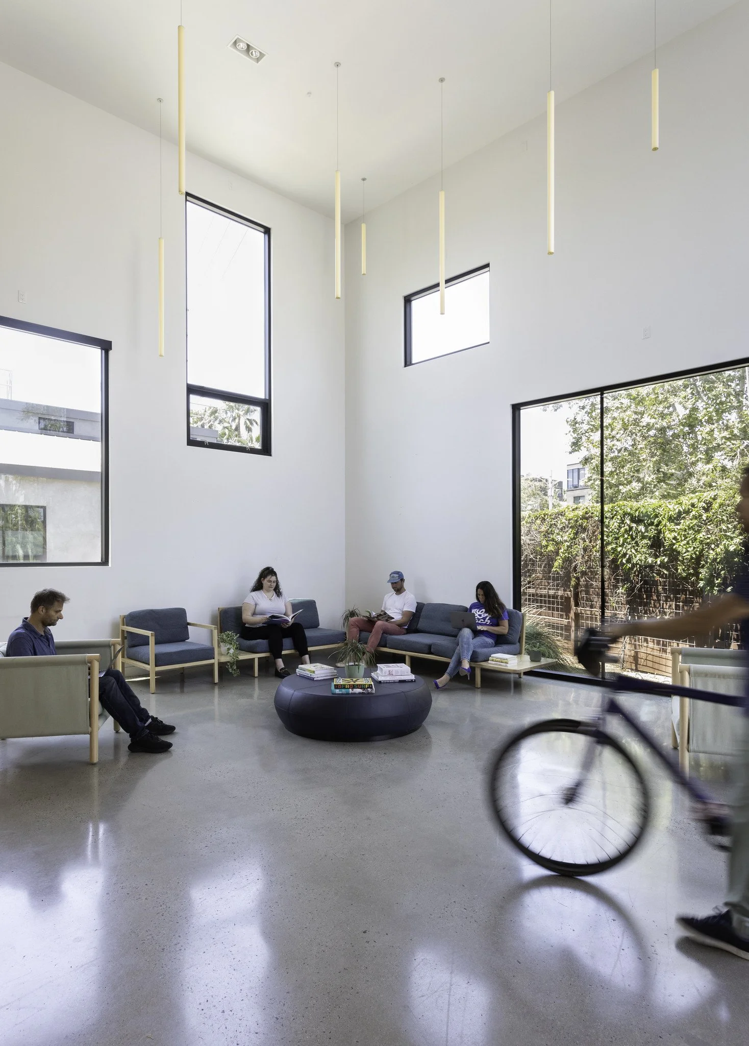Modern lounge or waiting area with high ceiling, white walls, large windows, and a large glass sliding door. There are five people sitting on couches, some reading or using laptops. A person on a bicycle is passing by in the foreground. The room feat