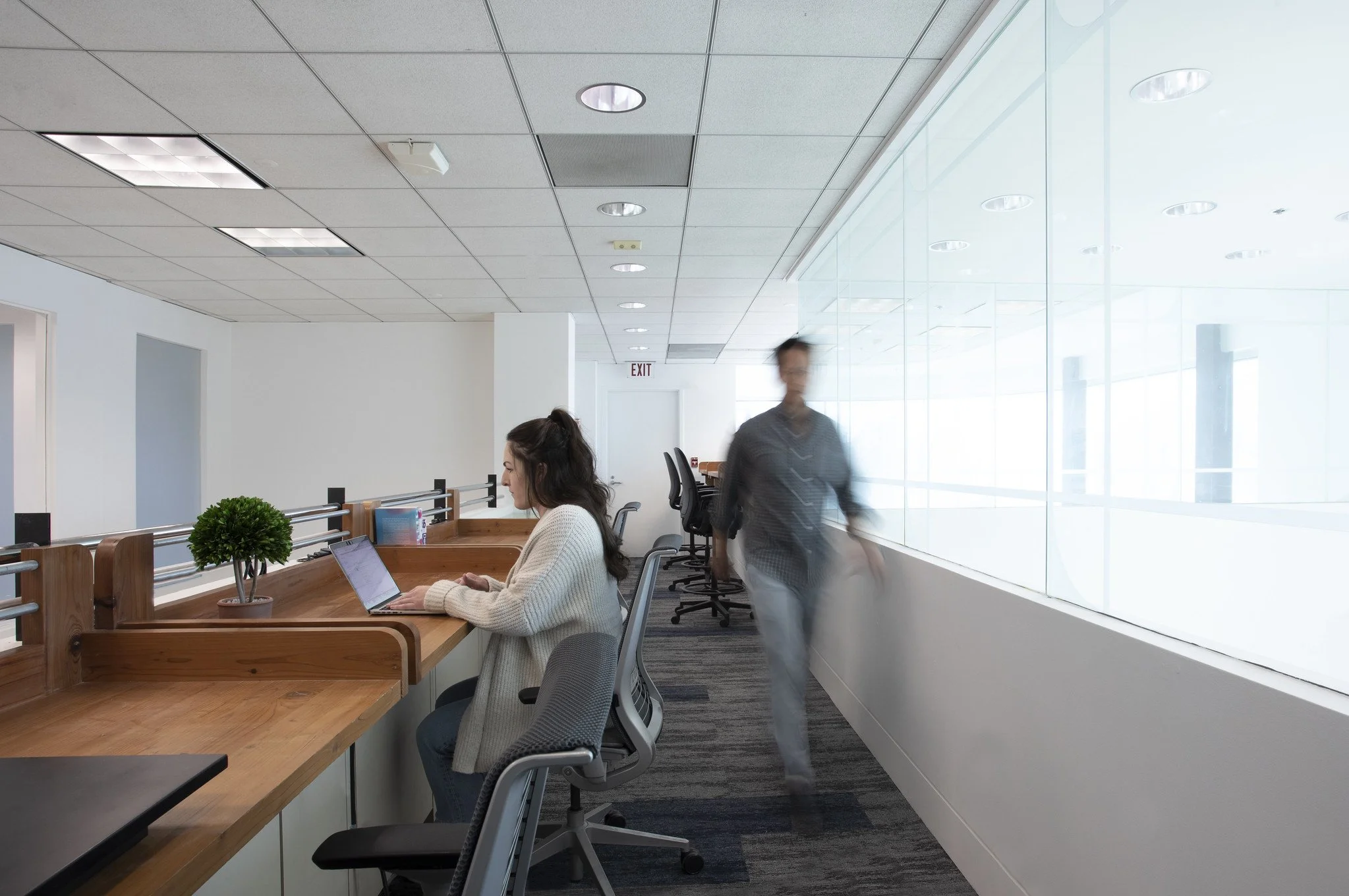 An office space with a woman working on a laptop at a wooden desk and a man walking past a large glass wall.