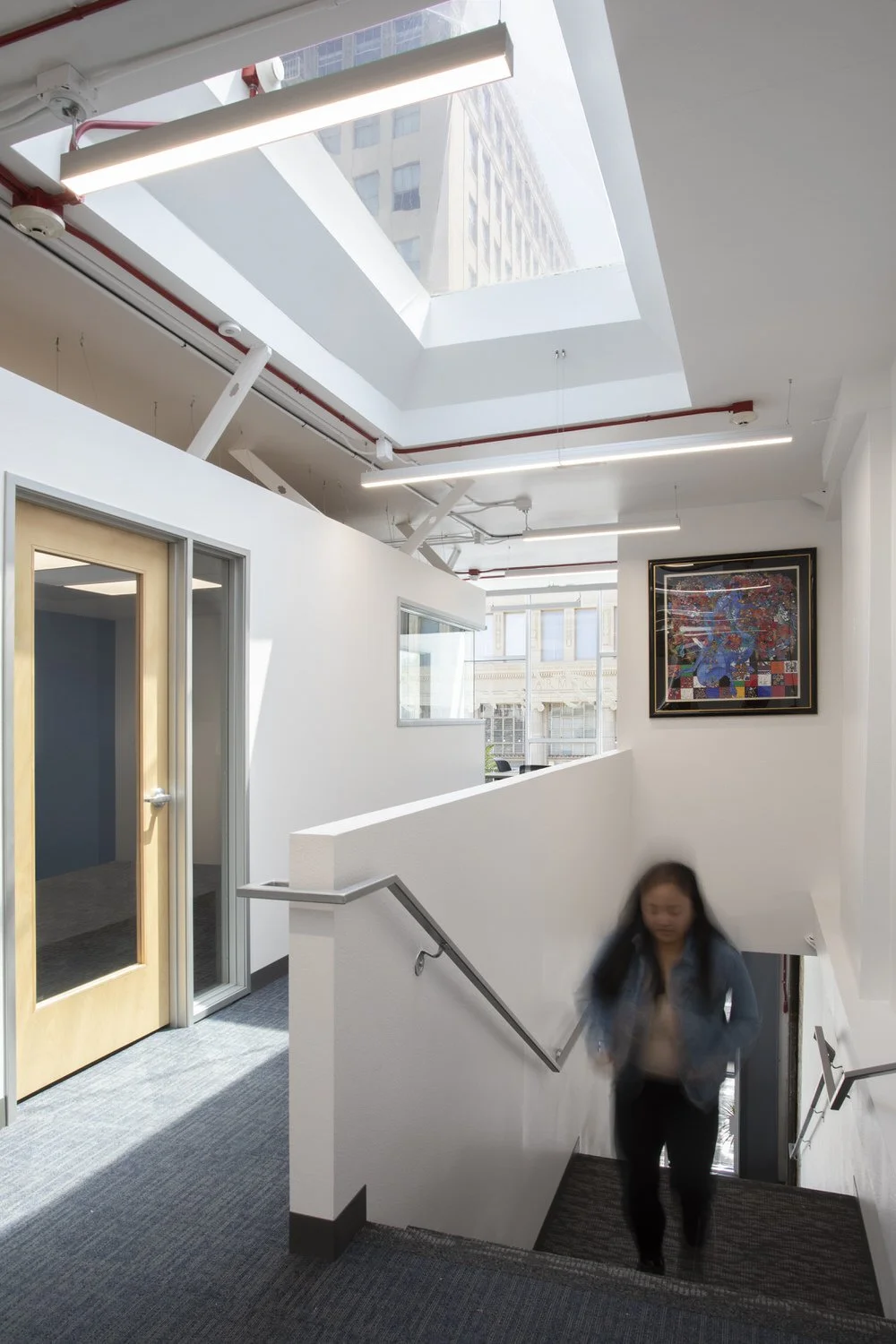 Office interior with staircase, large skylight, framed artwork, and a woman walking up stairs.