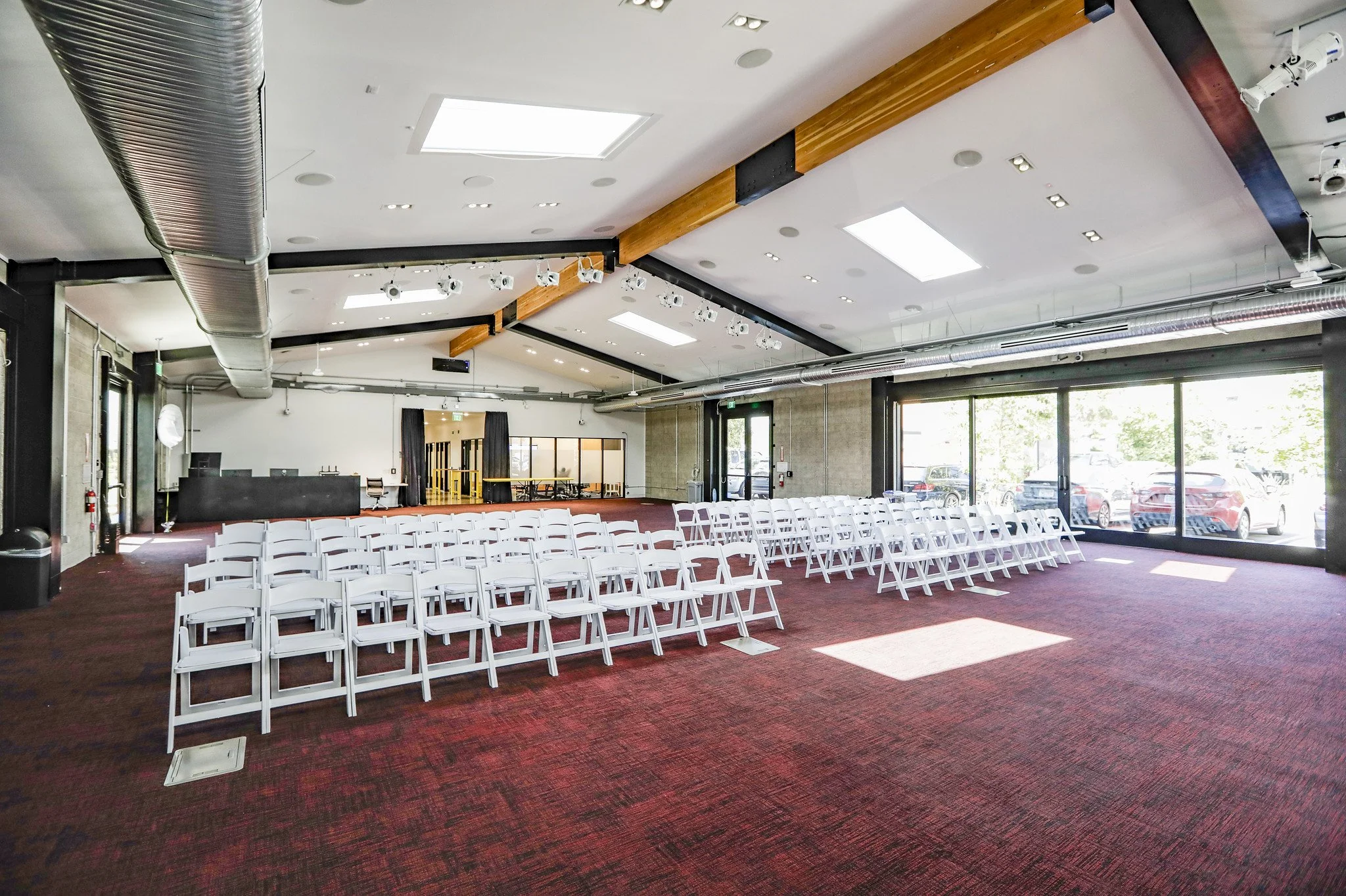 An empty event space with rows of white folding chairs facing a stage area. Large windows on one side showing parked cars outside. Ceiling with exposed ductwork and lighting fixtures. Red carpeted floor.