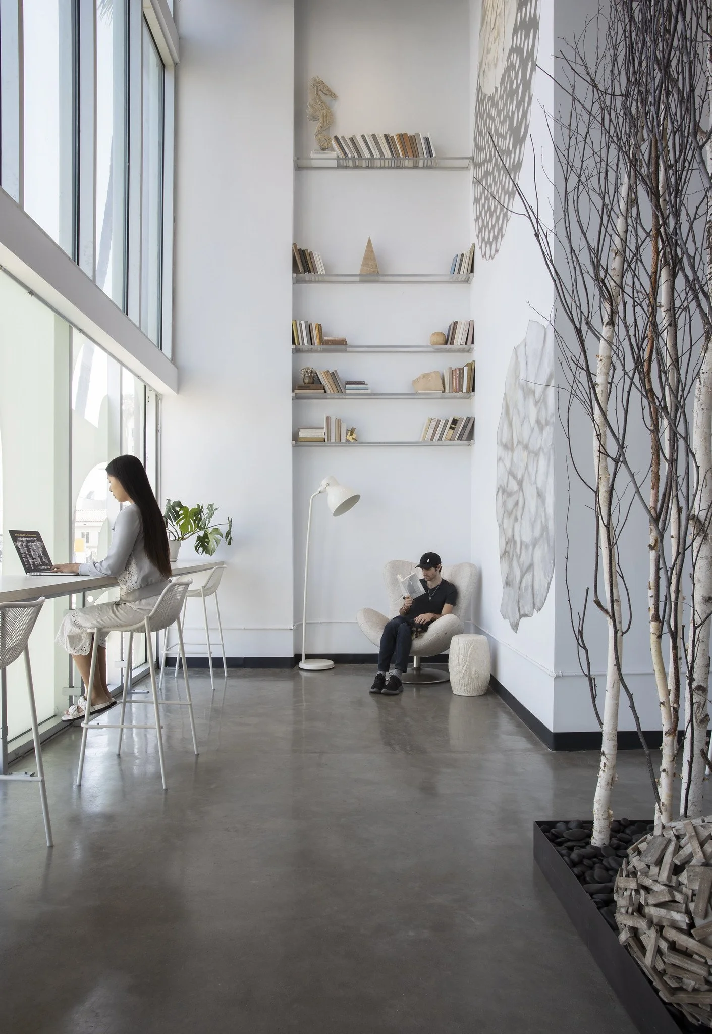 A modern indoor space with large windows, featuring a woman working at a high table by the window, and a man sitting in a cozy chair reading, with a tall white bookshelf on the wall filled with books and decorative items, and a corner with decorative