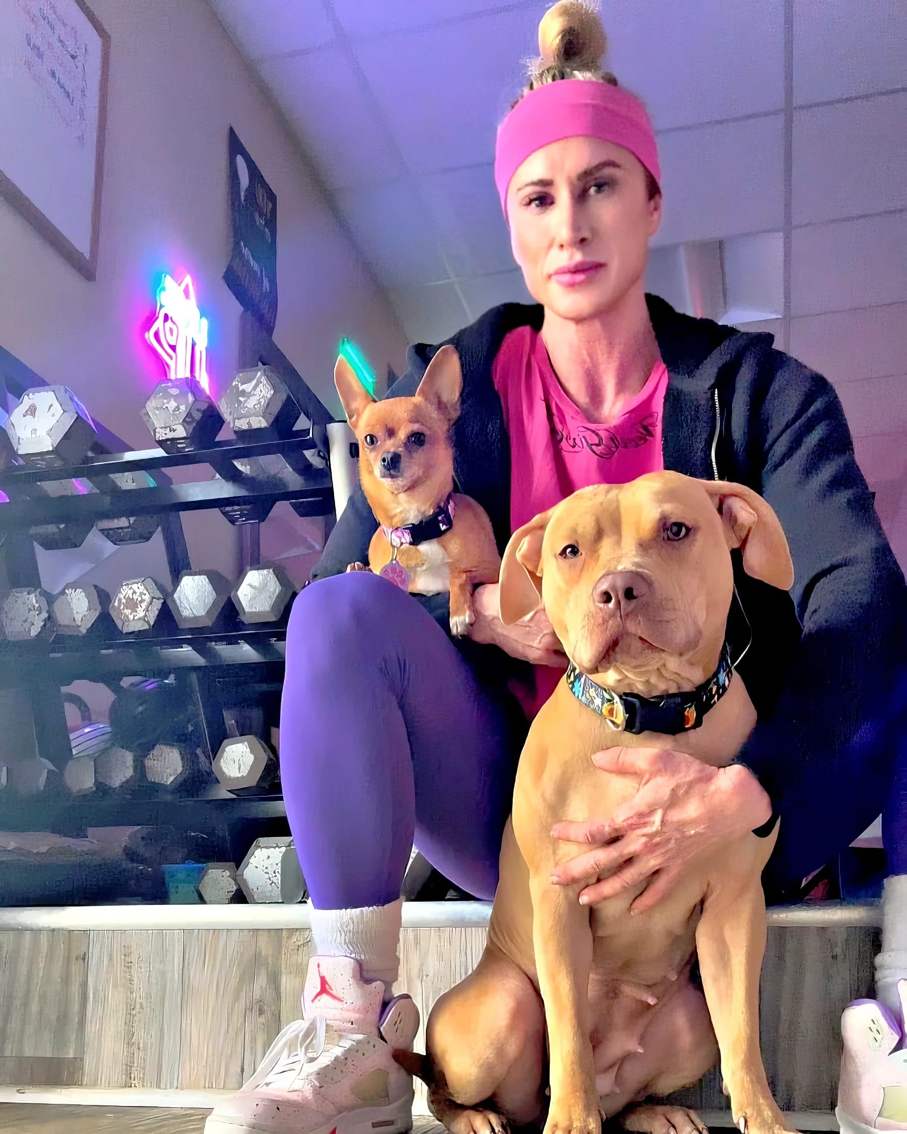A woman in a pink headband and purple leggings sitting with two dogs in a gym with neon lights and dumbbells in the background.