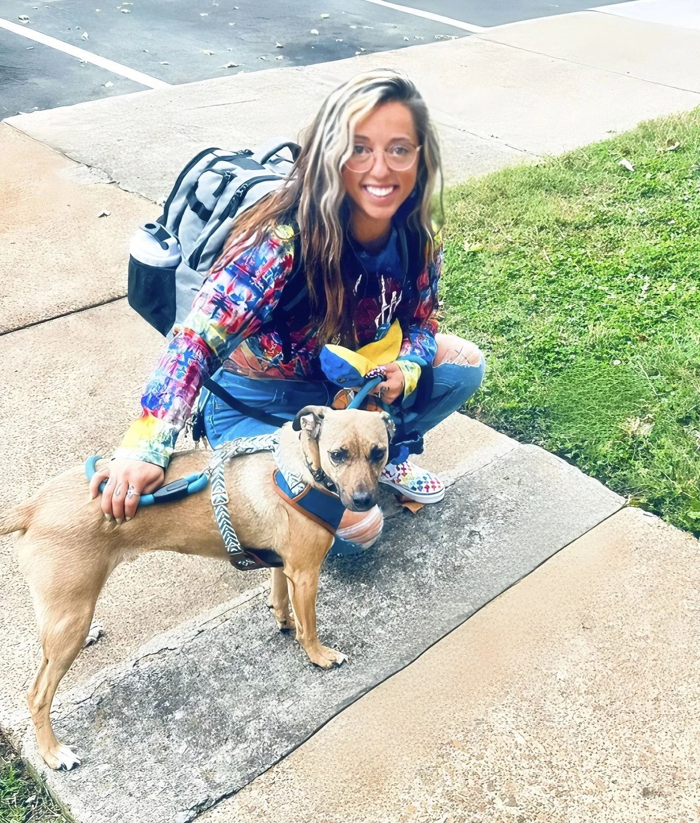 A smiling woman with glasses crouching beside a tan dog with a black nose on a leash outdoors on a sidewalk with grass and parking lot in the background.