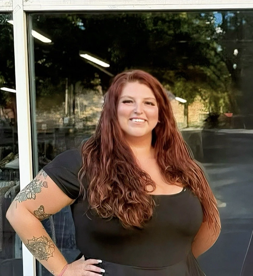 A woman with long, wavy red hair standing outside in front of a glass window.