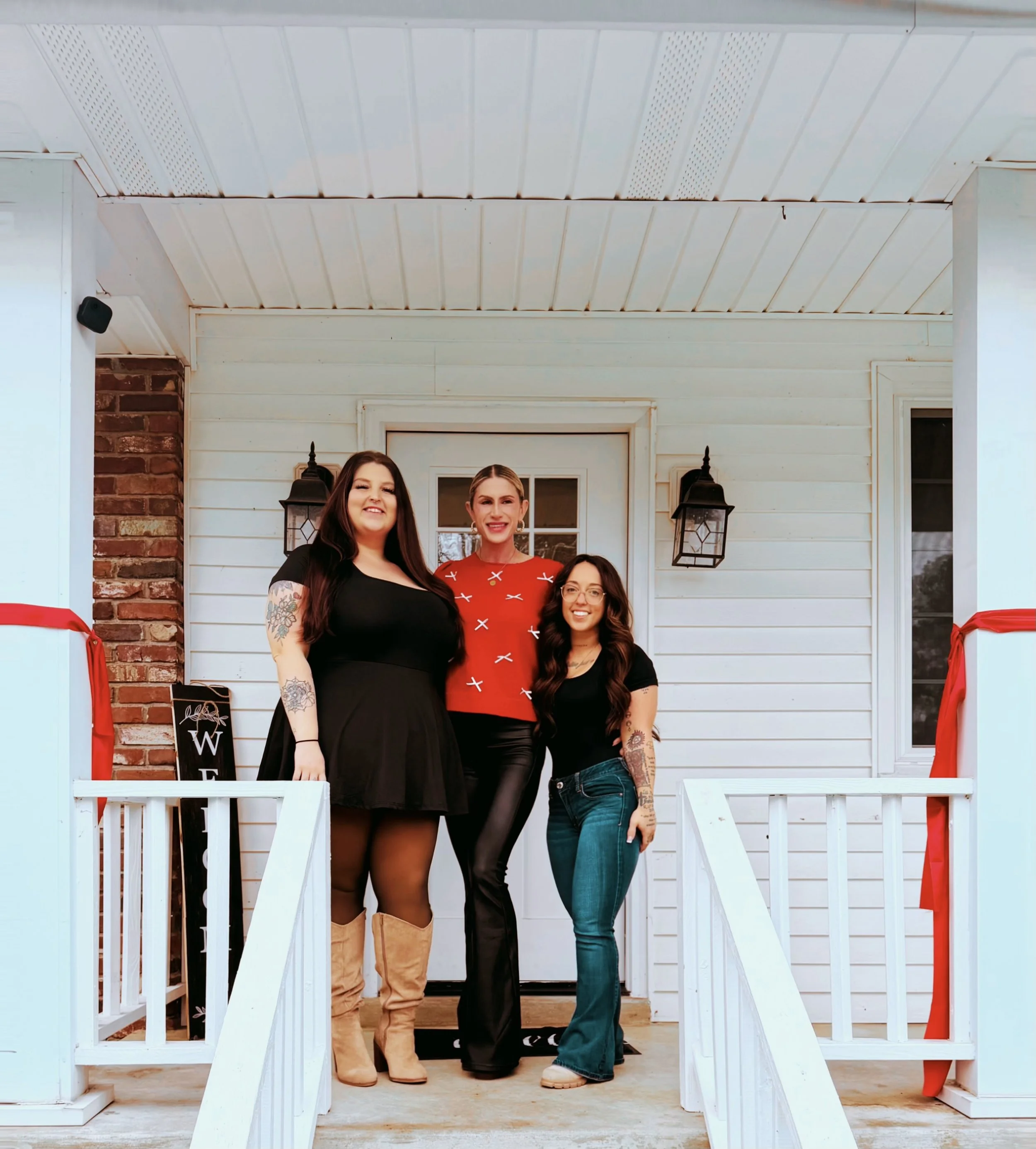 Three women standing on a porch in front of a white house with black lanterns, smiling at the camera. Two are wearing black tops and jeans, and one is wearing a red top with black pants. There are red ribbons on both sides of the porch.