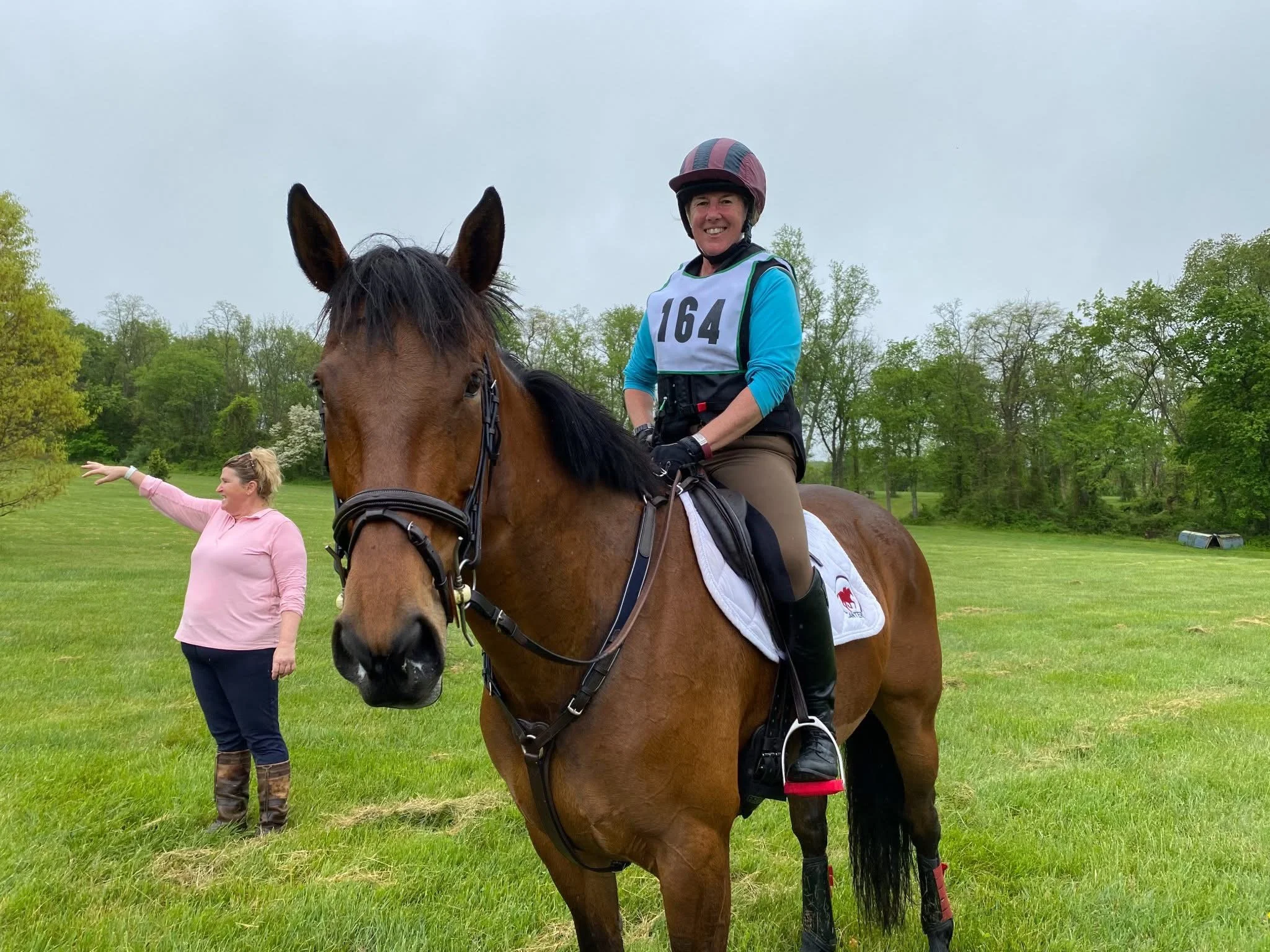 A woman in riding gear with a helmet and a bib numbered 164, sitting on a brown horse in a green field. A woman in a pink sweater and boots stands nearby, gesturing with her arm.