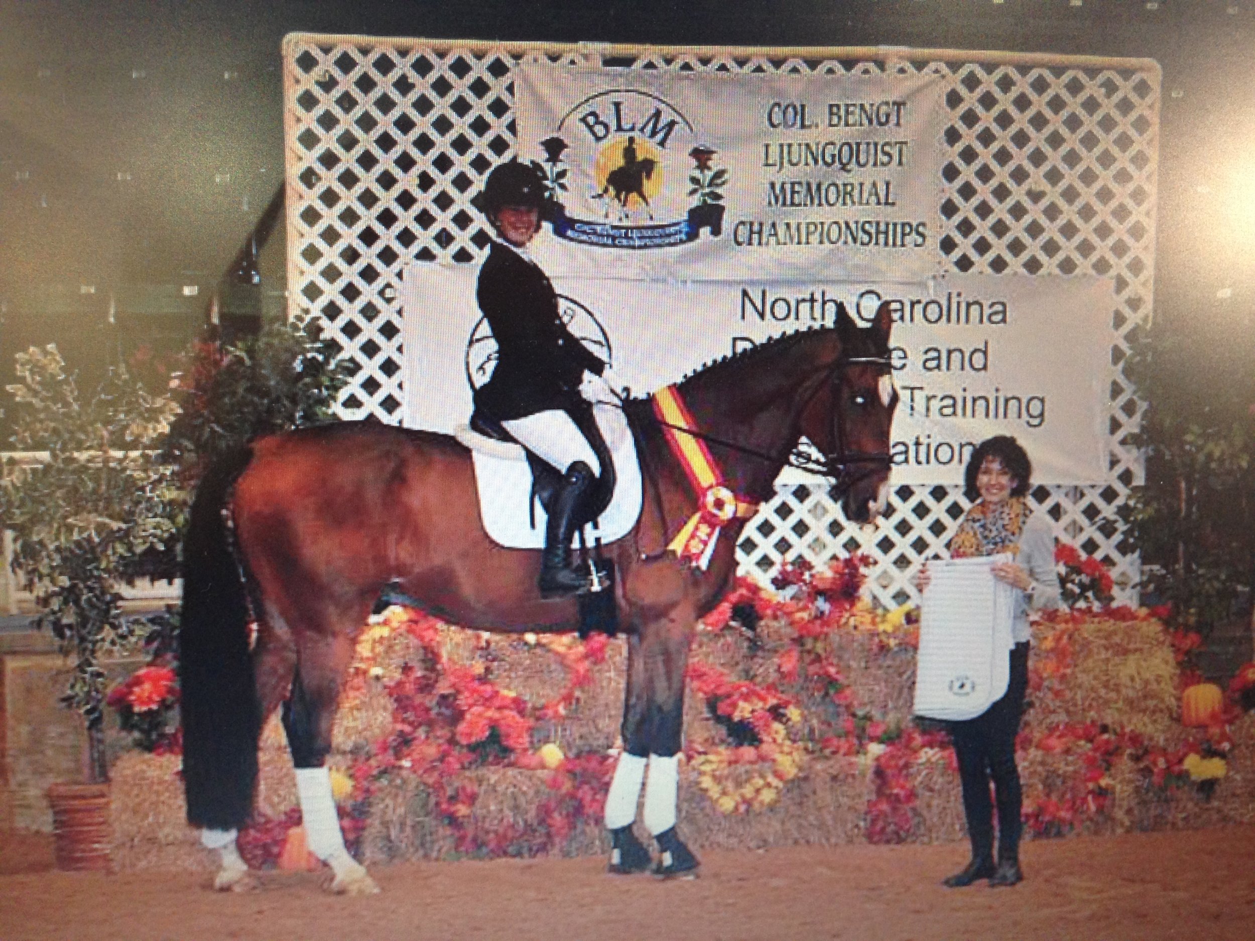 A woman on horseback receiving an award at an equestrian event, with a woman presenting a ribbon. The background features a banner and fall decorations.