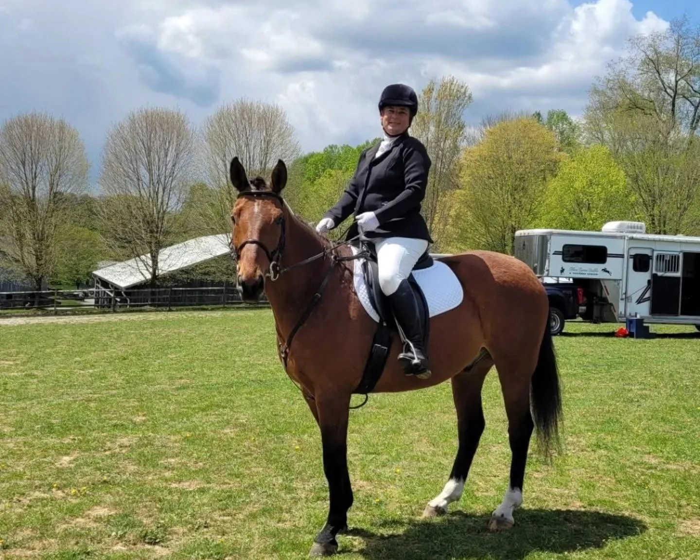 A woman dressed in equestrian attire sitting on a brown horse in an outdoor field, with horse trailers and trees in the background.