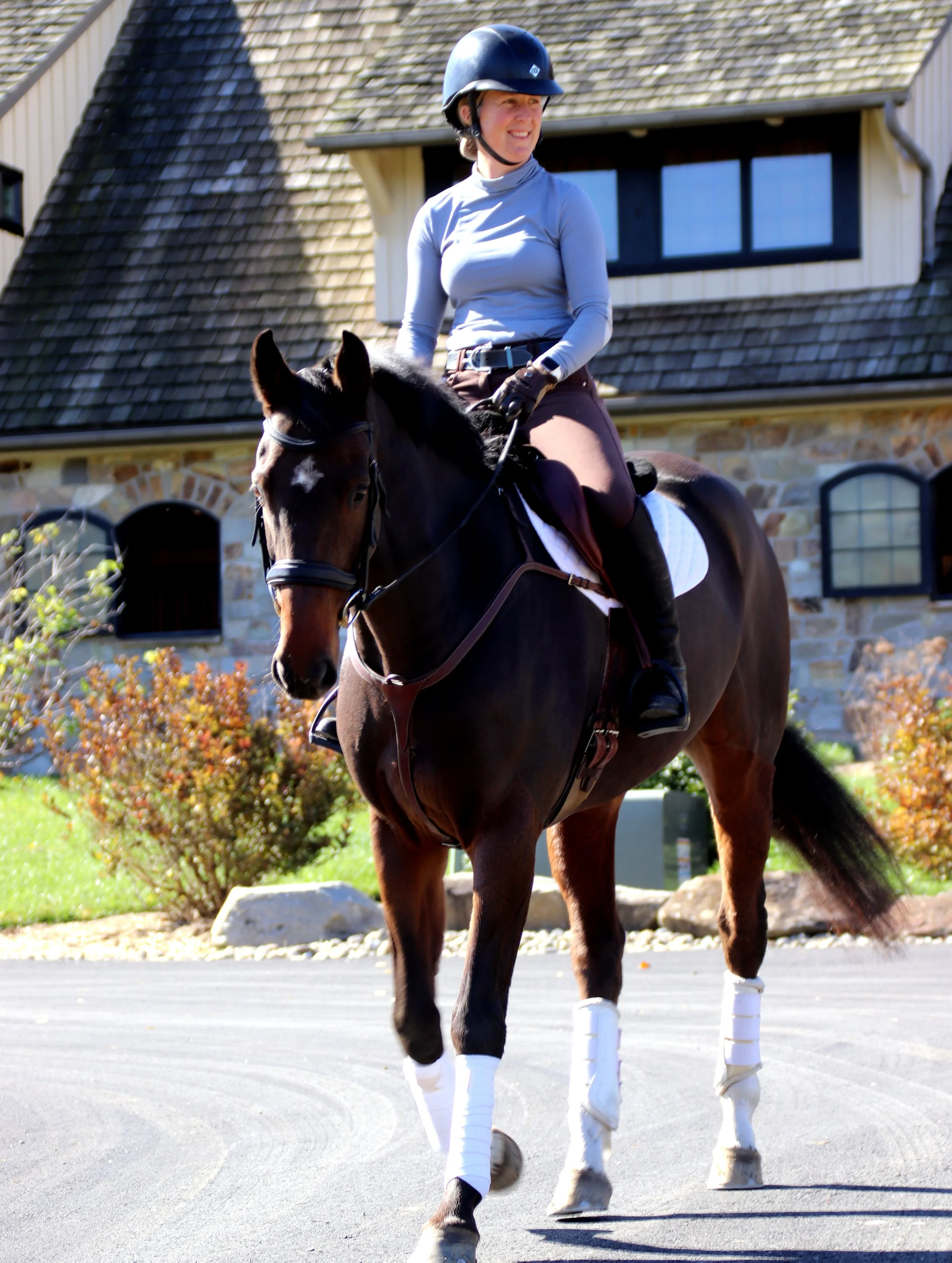 A woman riding a dark brown horse outdoors in front of a house with stone walls and shingled roof, wearing a helmet, light blue long-sleeve shirt, and brown riding pants.