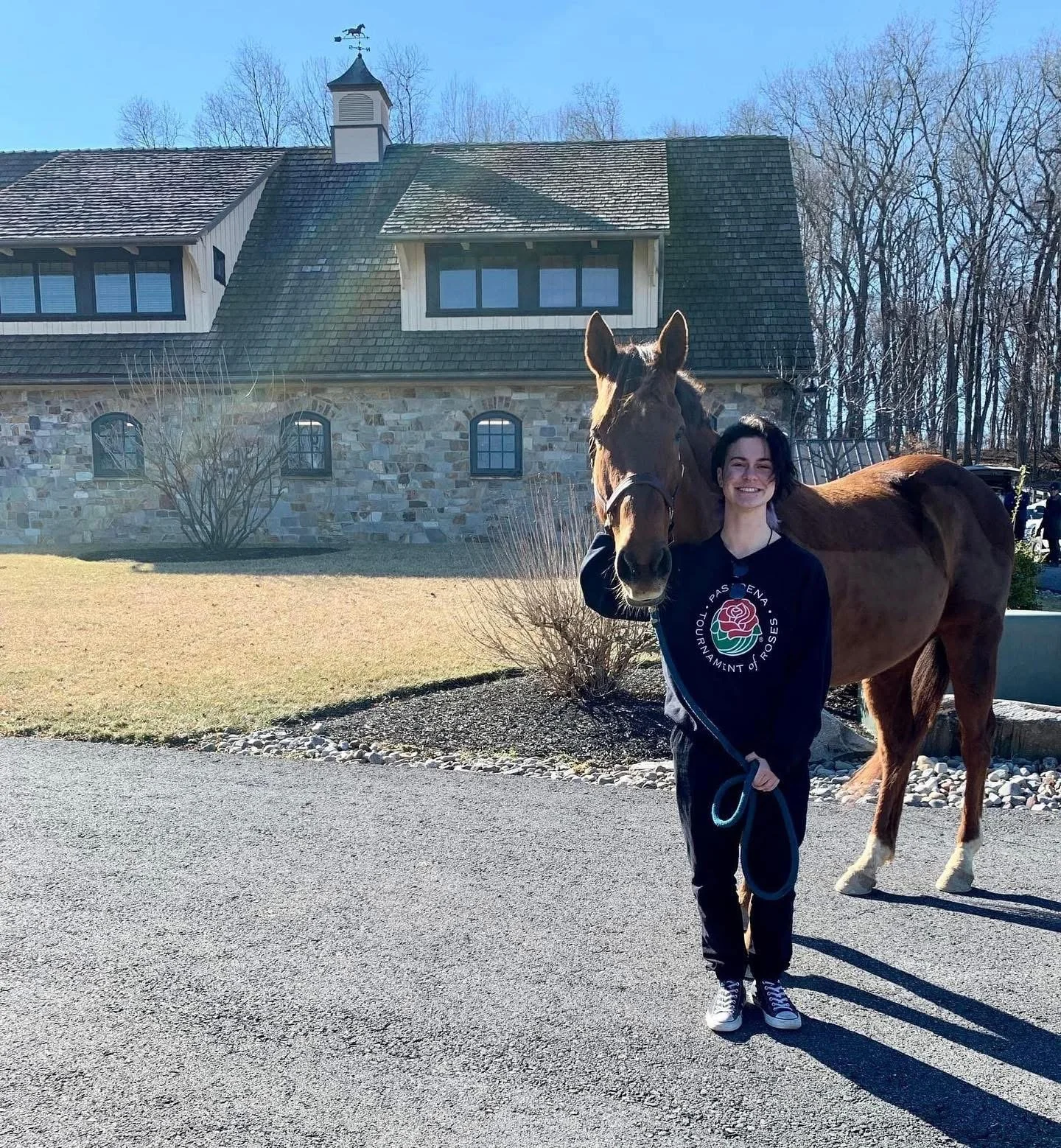 A woman standing outside holding a brown horse's lead rope, smiling, with a house and leafless trees in the background.