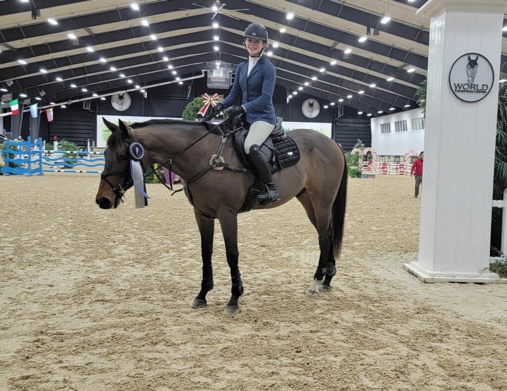 A woman in equestrian attire sitting on a brown horse inside an indoor riding arena, holding a ribbon, indicating she won or placed in a competition.