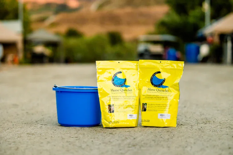 Two yellow bags of horse quincher supplement and a blue bucket outdoors on a gravel surface with blurred background of tents and hills.