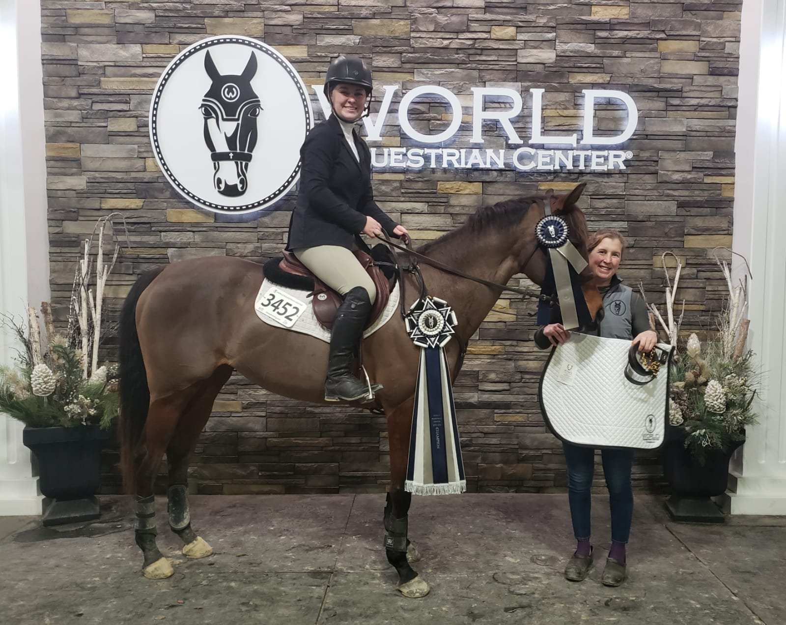 A woman on a horse holding a ribbon, and another woman standing next to the horse holding a white saddle pad, celebrating at the World Equestrian Center.