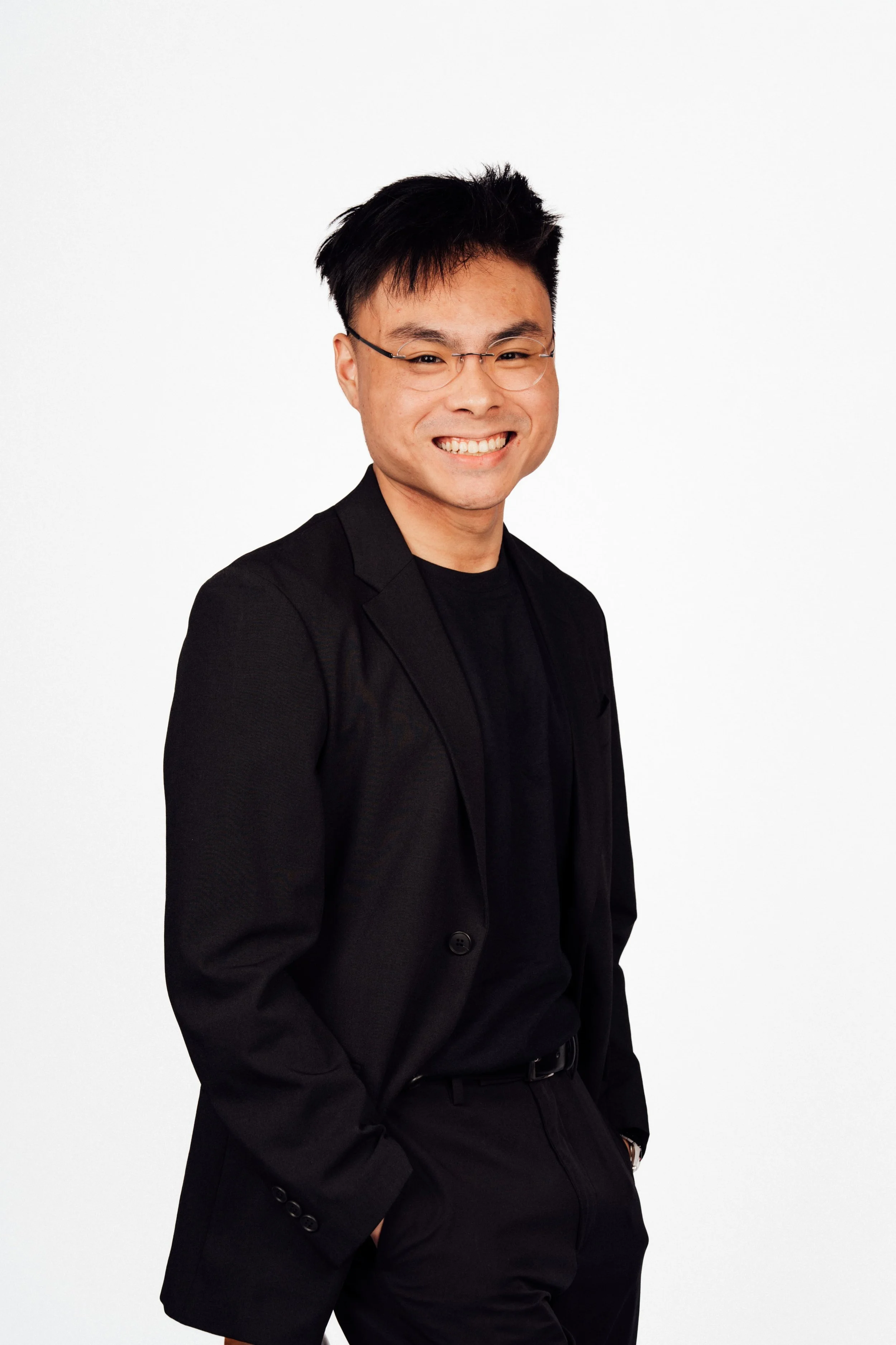 A man with glasses and short black hair wearing a black blazer and black t-shirt, smiling with hands in pockets against a plain white background.