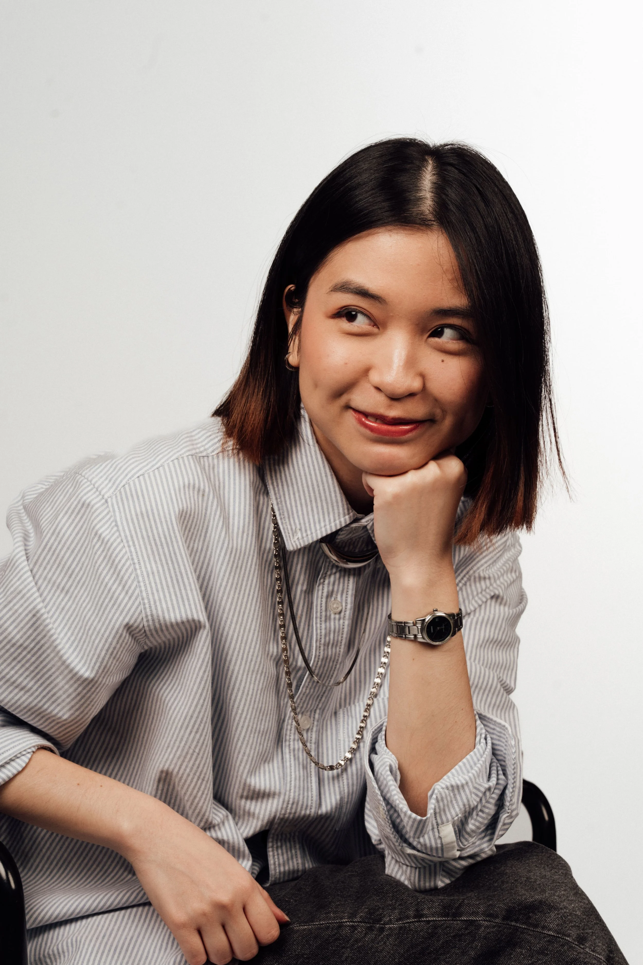 A young woman with shoulder-length dark hair, wearing a striped button-up shirt, sitting on a chair with her chin resting on her hand, looking to the side, smiling subtly, against a plain white background.
