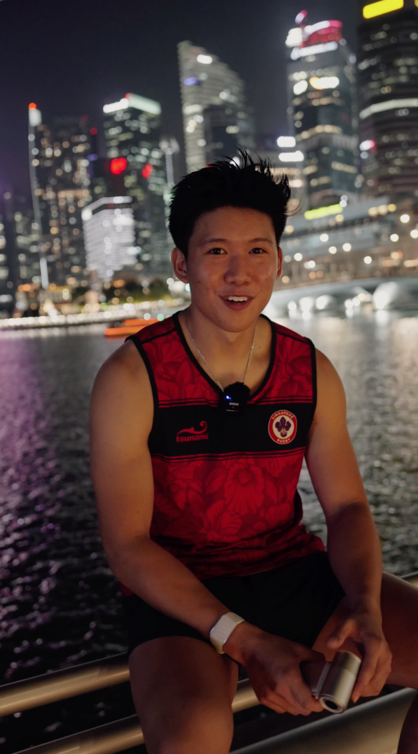 A young man wearing a red and black sports jersey with a Singapore Lion crest, sitting outdoors at night by a body of water with a city skyline illuminated in the background.