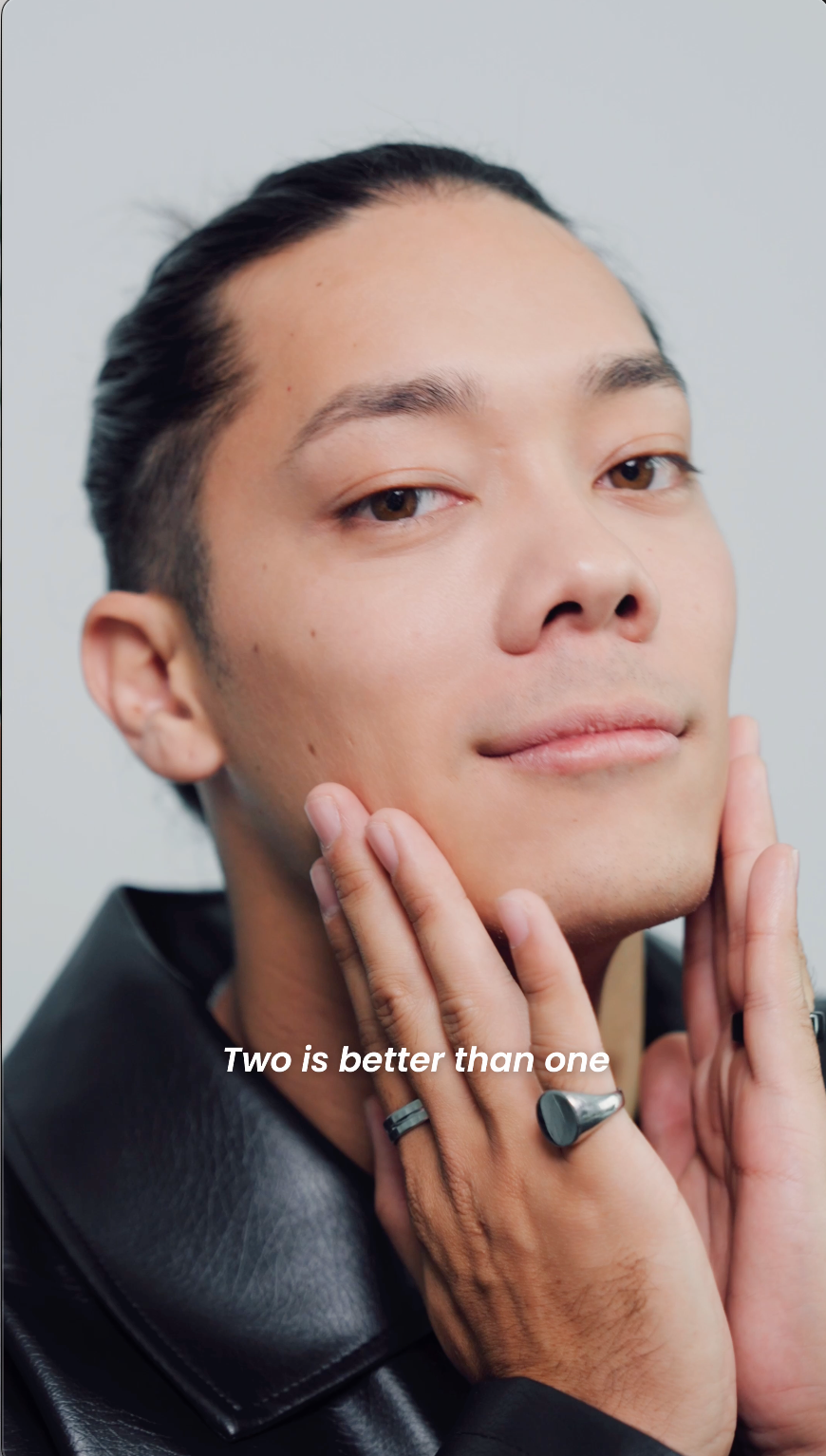 Close-up of a person with slicked-back hair and smooth skin, touching their face with both hands. They are wearing a black leather jacket and rings, with the caption, "Two is better than one."