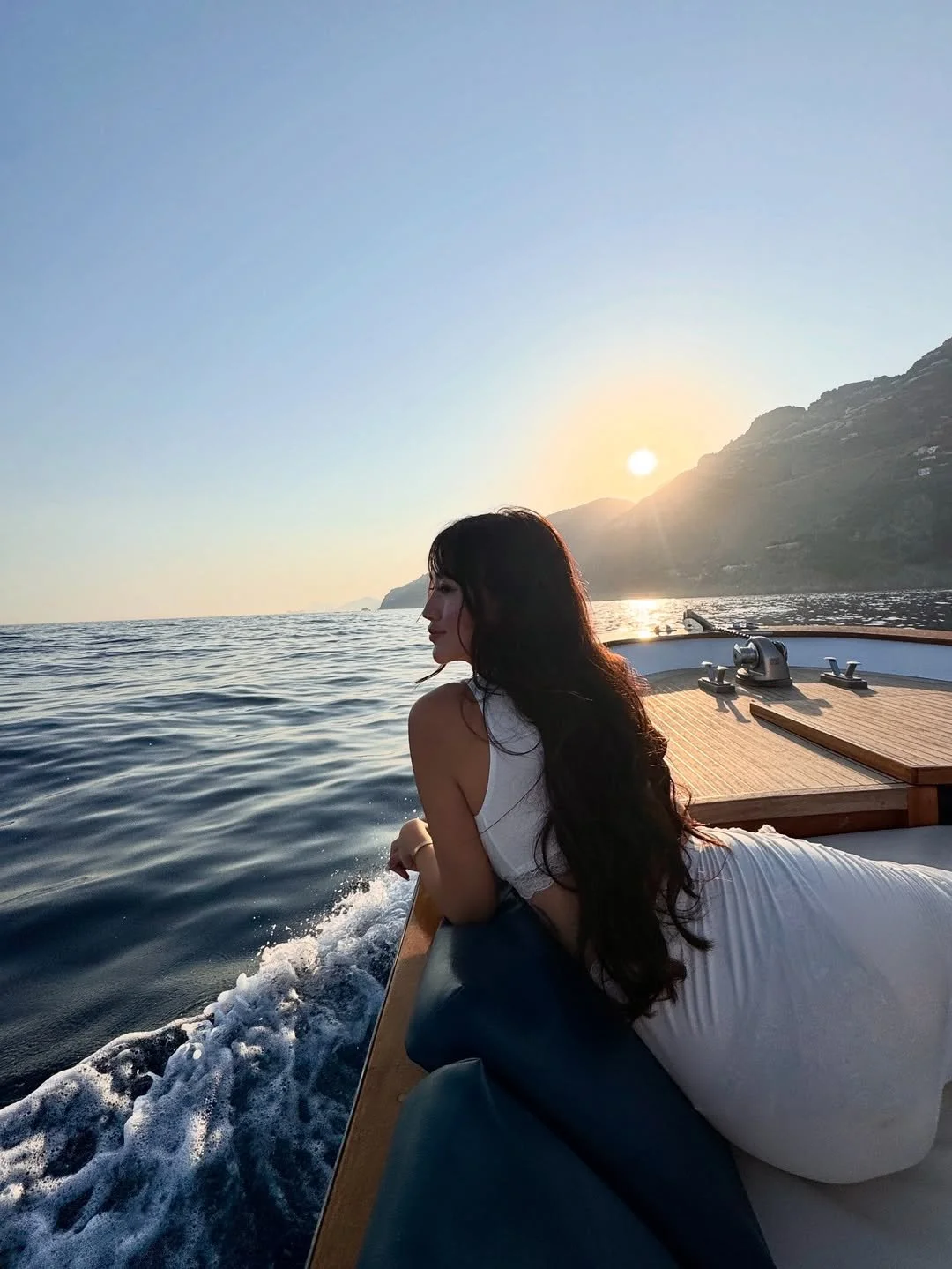 A woman with long dark hair relaxing on a boat during sunset, overlooking the ocean with mountains in the background.