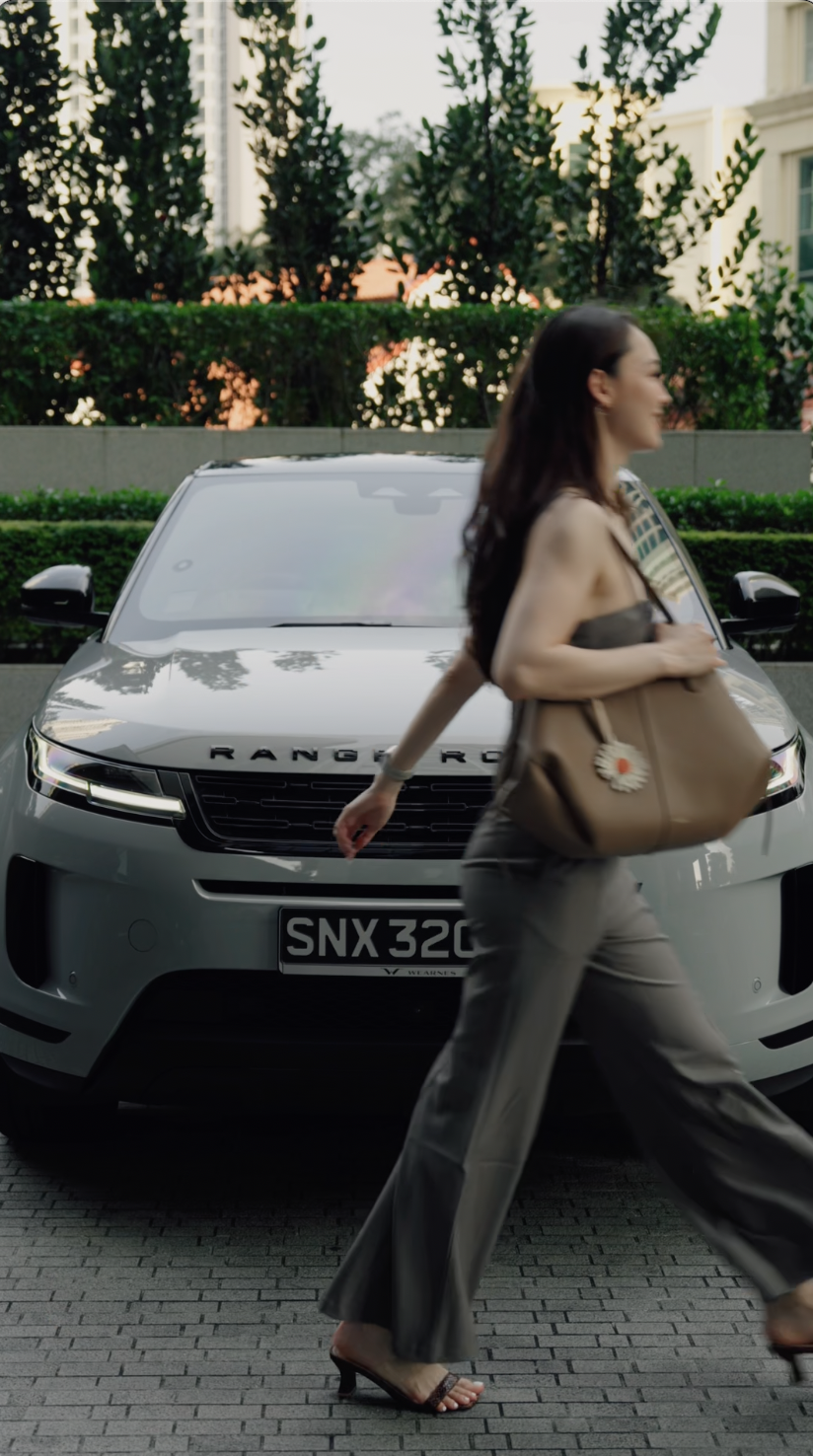 A woman in a beige strapless top and gray wide-leg pants walking past a silver Range Rover with a black grille, in an outdoor setting with greenery and tall trees in the background.