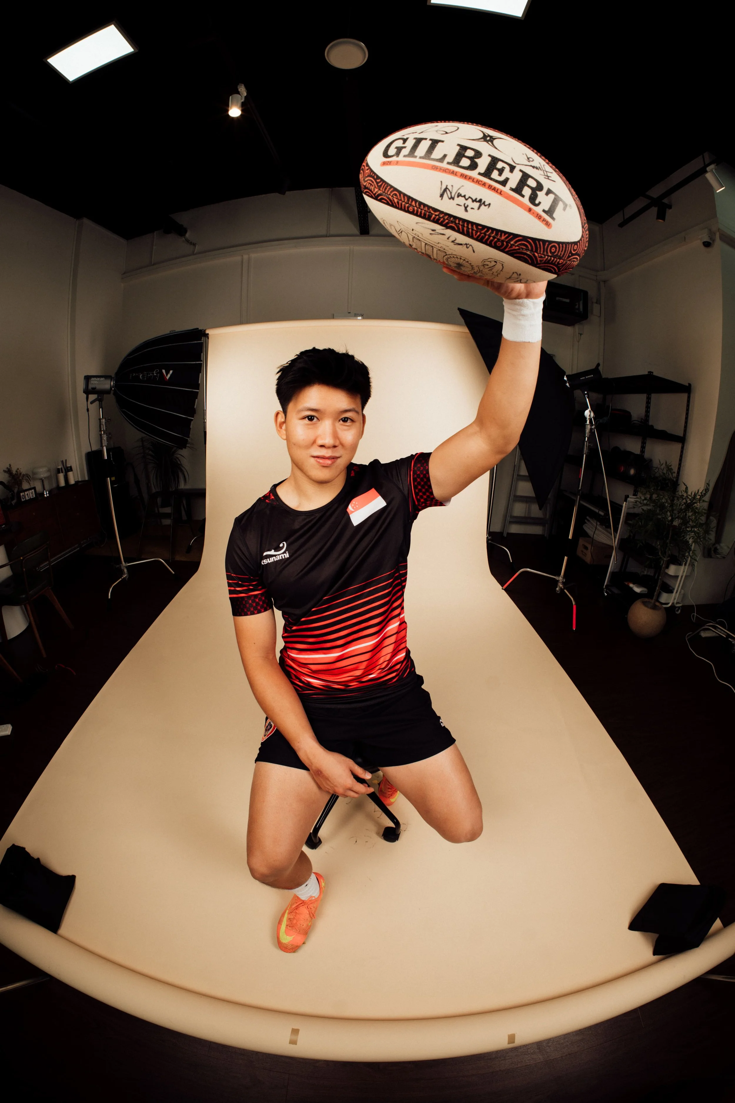 A young man in sportswear kneeling on one knee, holding an Australian football above his head in a photography studio.