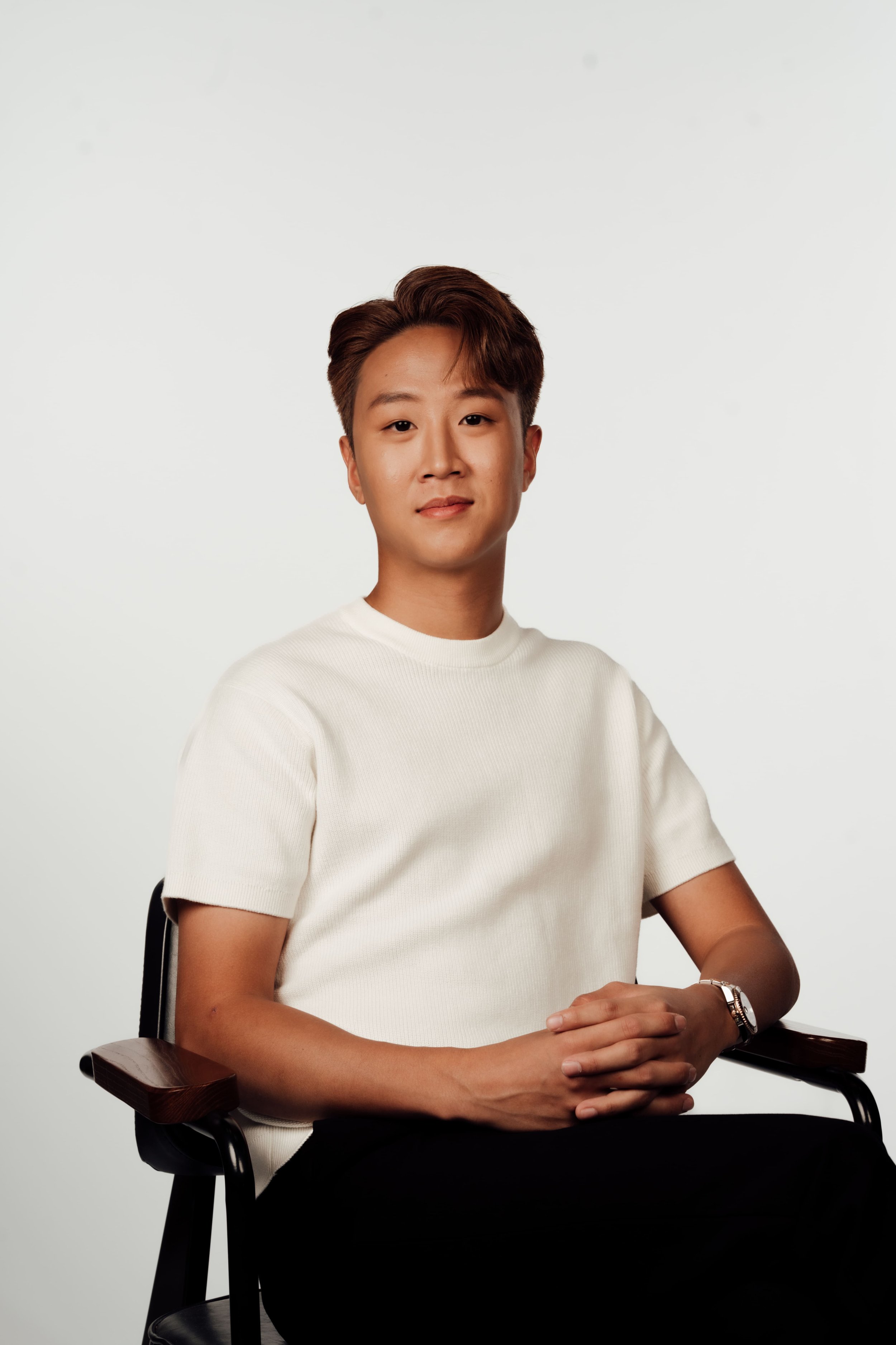 A young man with short, styled hair sitting on a black chair in front of a plain white background. He is wearing a white t-shirt and a wristwatch, and he has a calm, confident expression.