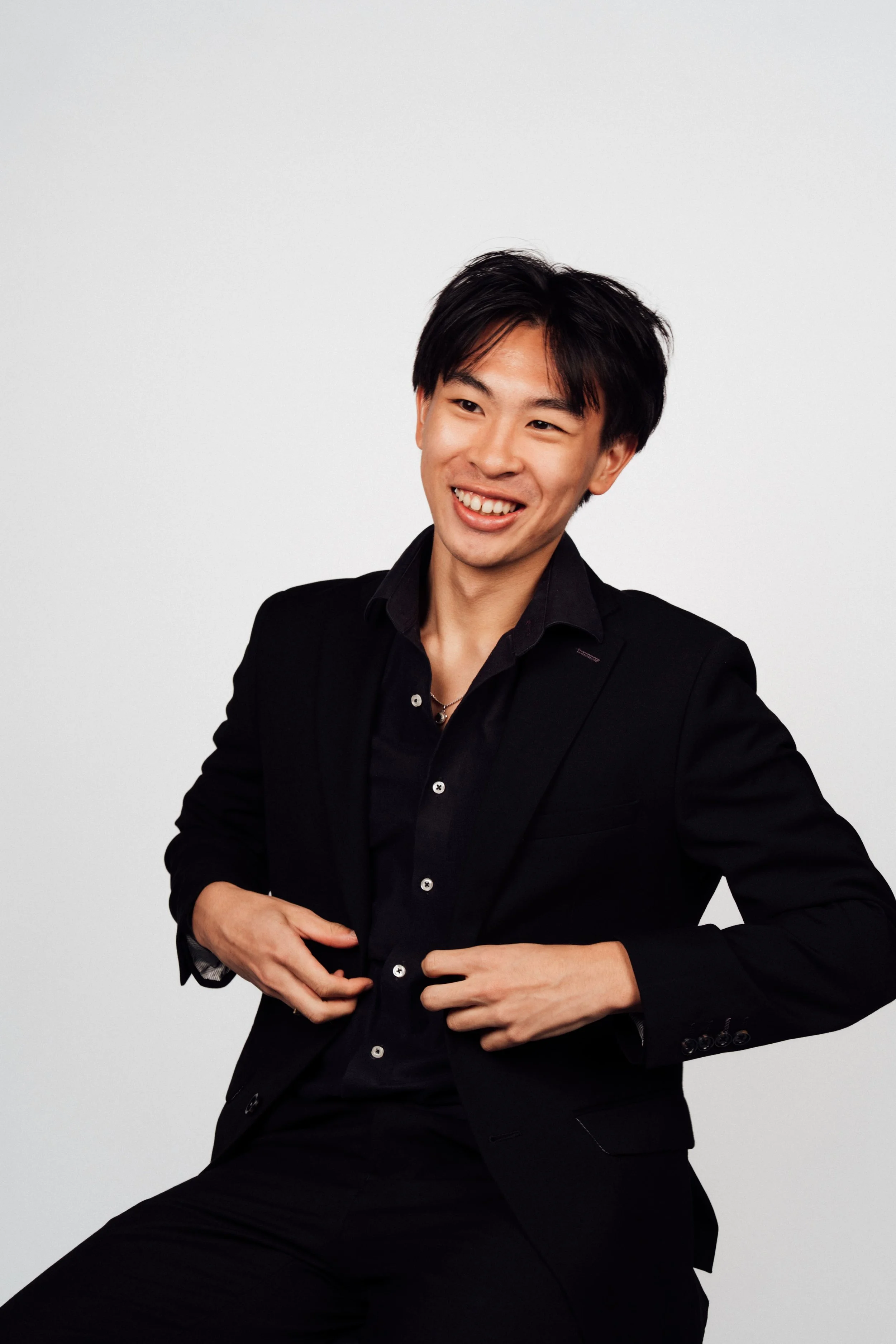 A young man with black hair wearing a black blazer and shirt, smiling, against a plain white background.