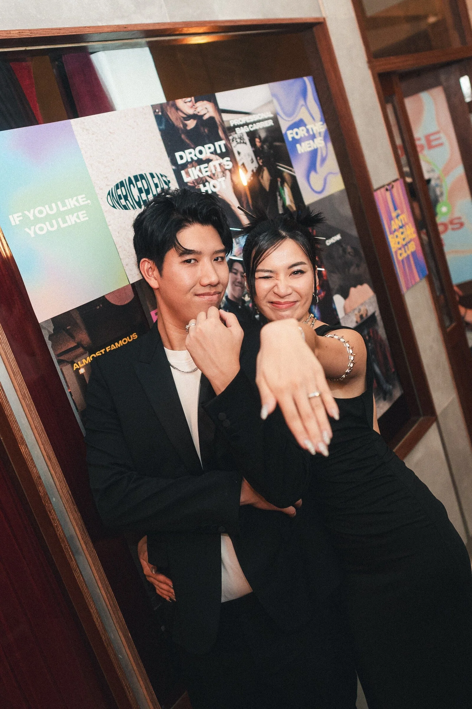 Two young adults, a man and a woman, posing playfully in front of a colorful wall with posters. The woman is showing off her ring and smiling, while the man has his arm crossed and is looking at the camera.