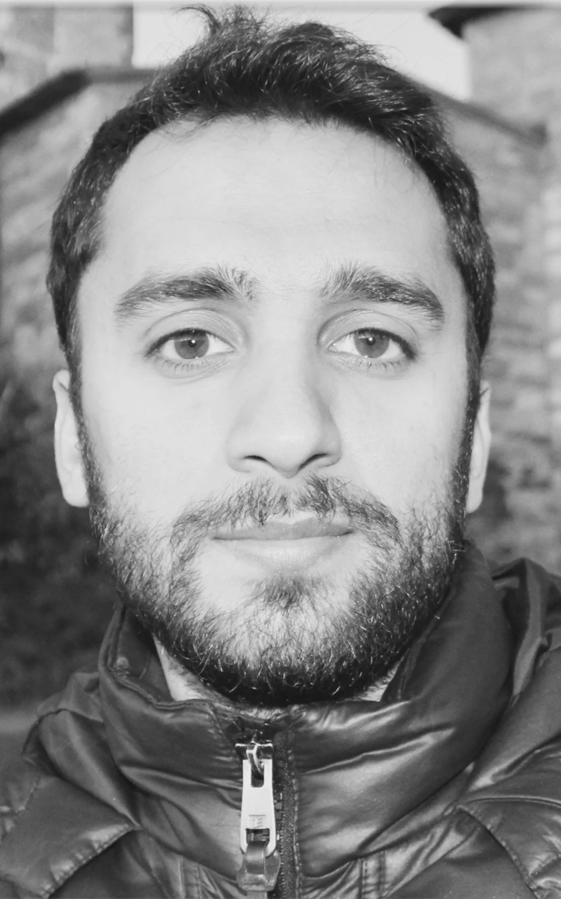 Et svart-hvitt portrett av en mann med kort, mørkt hår, skjegg og kraftige øyenbryn, iført en tykk jakke med glidelås. Bakgrunnen er uklar.