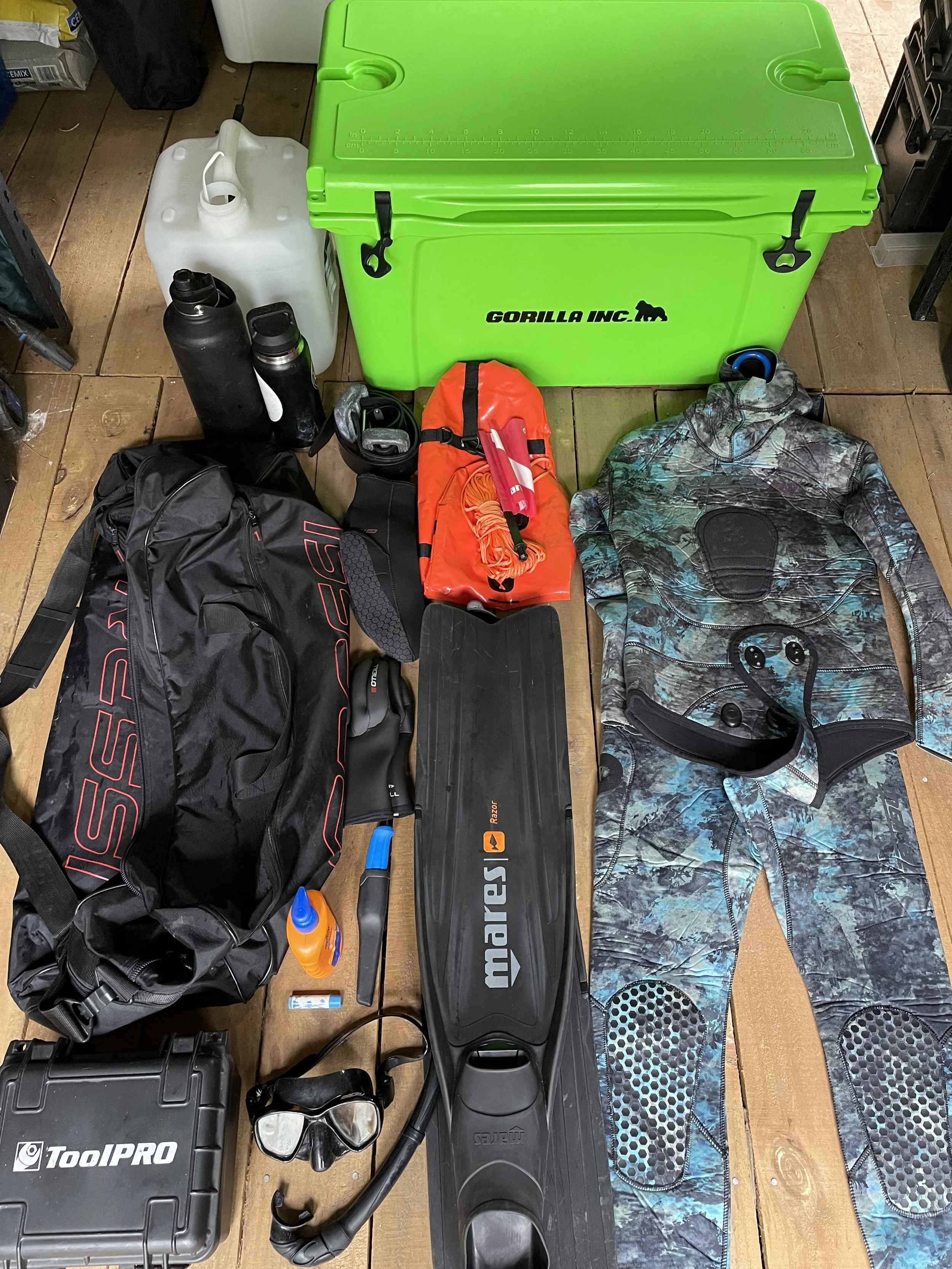 Scuba diving or snorkeling gear including wetsuit, fins, snorkel, mask, 2 water bottles, a large green cooler, black toolbox, and other accessories arranged on a wooden floor.