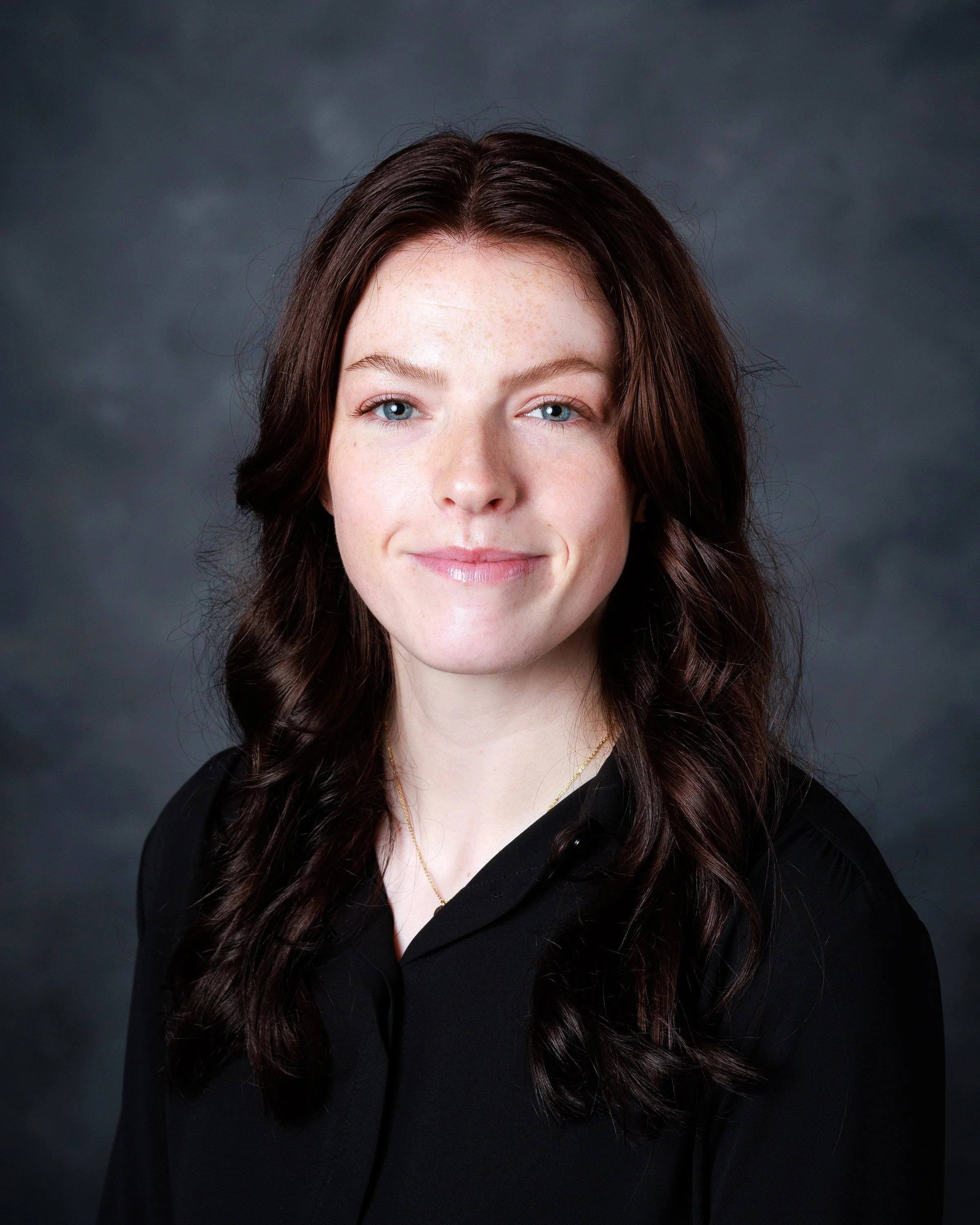 A woman with long dark hair and blue eyes smiling at the camera against a dark gray background.