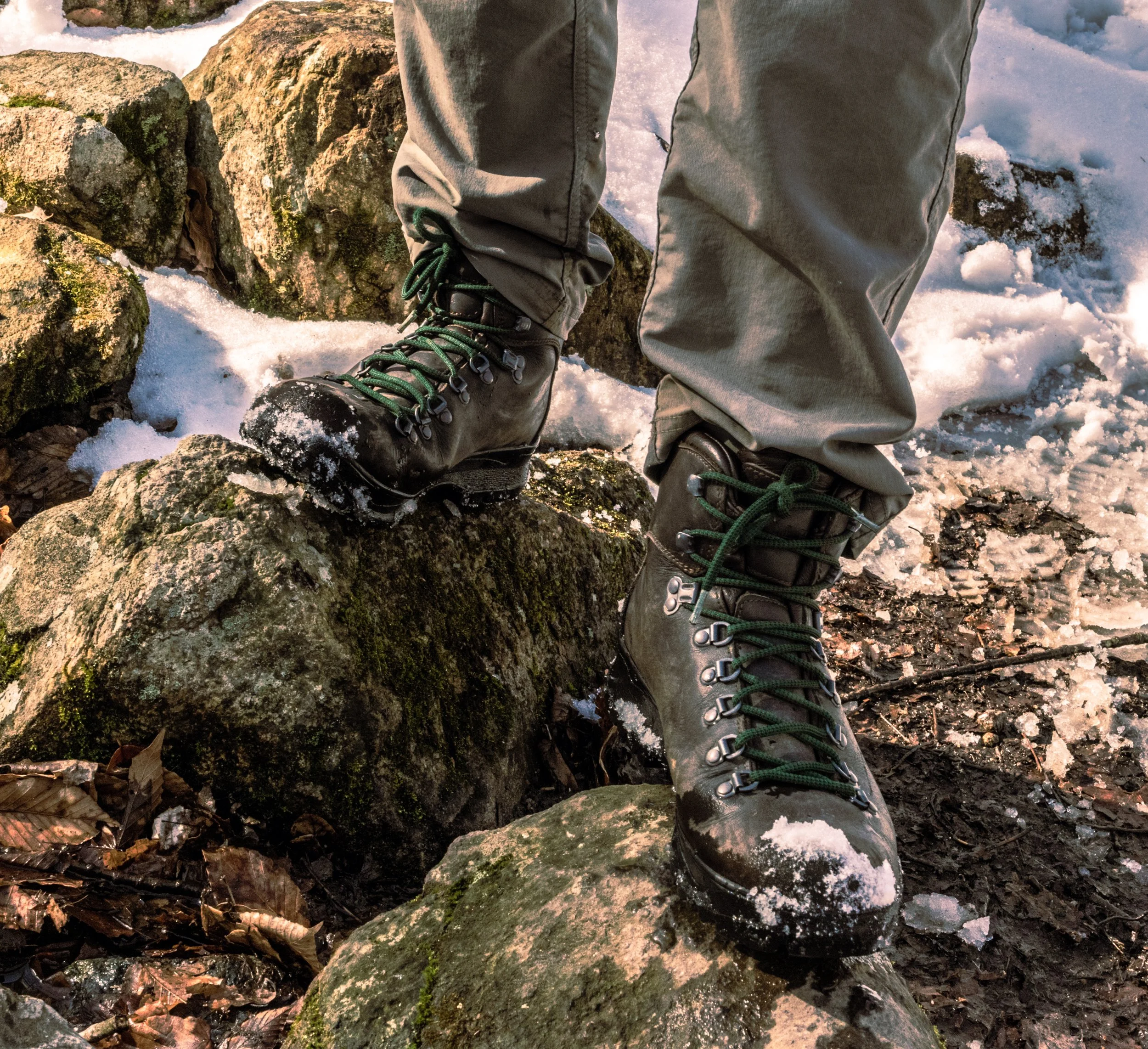 Close-up of a person wearing black hiking boots and khaki pants navigating on moss-covered rocks in a snowy outdoor setting.