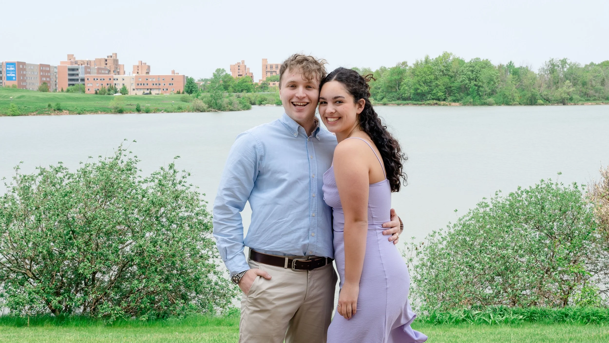A young couple standing close together smiling near a river with greenery and buildings in the background.