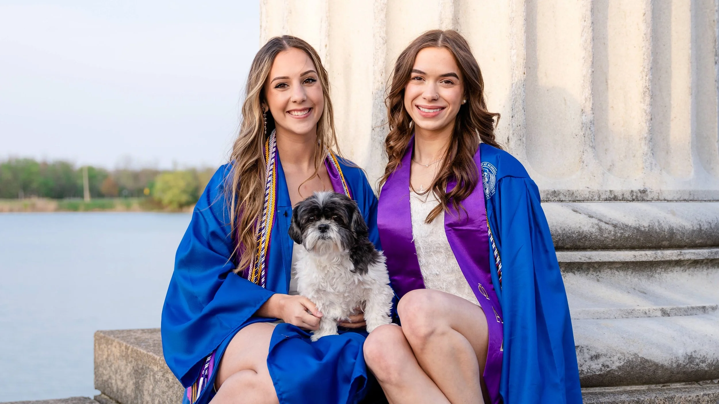 Two young women in graduation gowns sitting outdoors near a water body with a small dog between them.