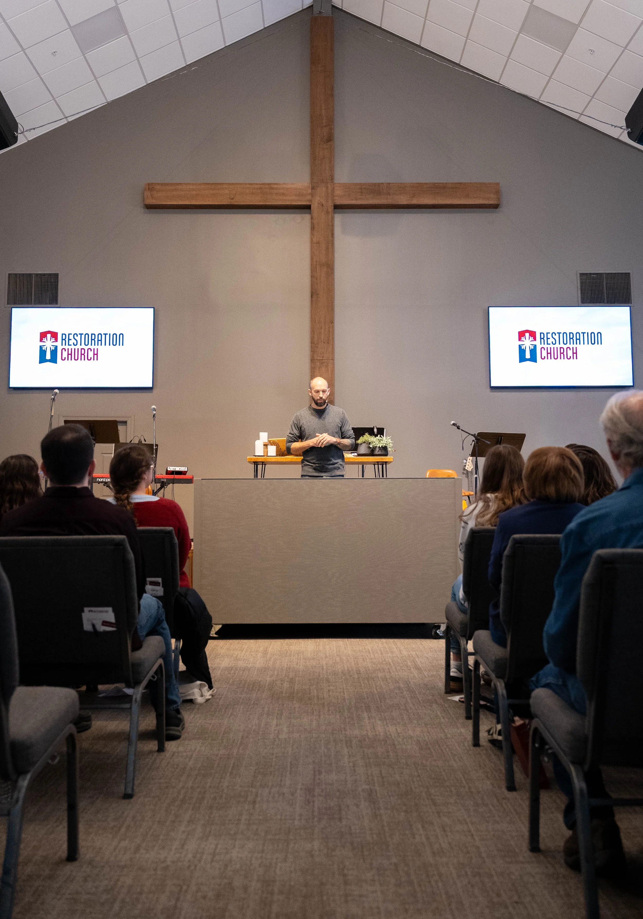 A man speaking at a church service in front of a wooden cross on the wall, with two screens displaying 'Restoration Church' logos, and an audience seated facing the stage.