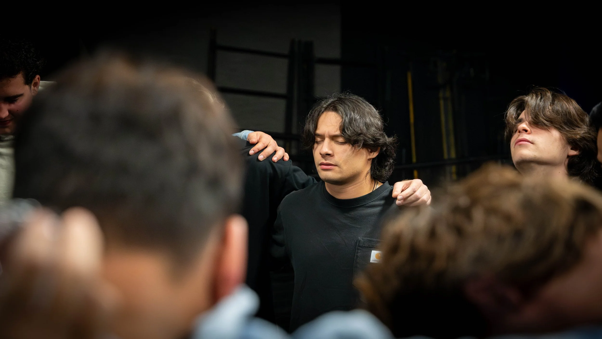 A man with black hair, eyes closed, appears to be praying or meditating, with his arms resting on others' shoulders in a group.