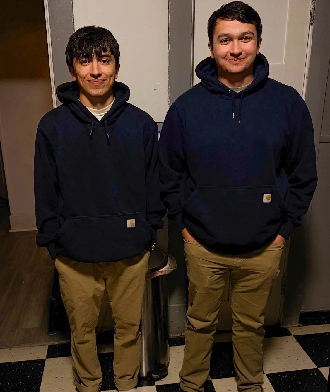 Two men standing side by side indoors, both wearing dark hoodies and khaki pants. The man on the left has dark hair with bangs, and the man on the right has short, dark hair. They are smiling and have their hands in their pockets.