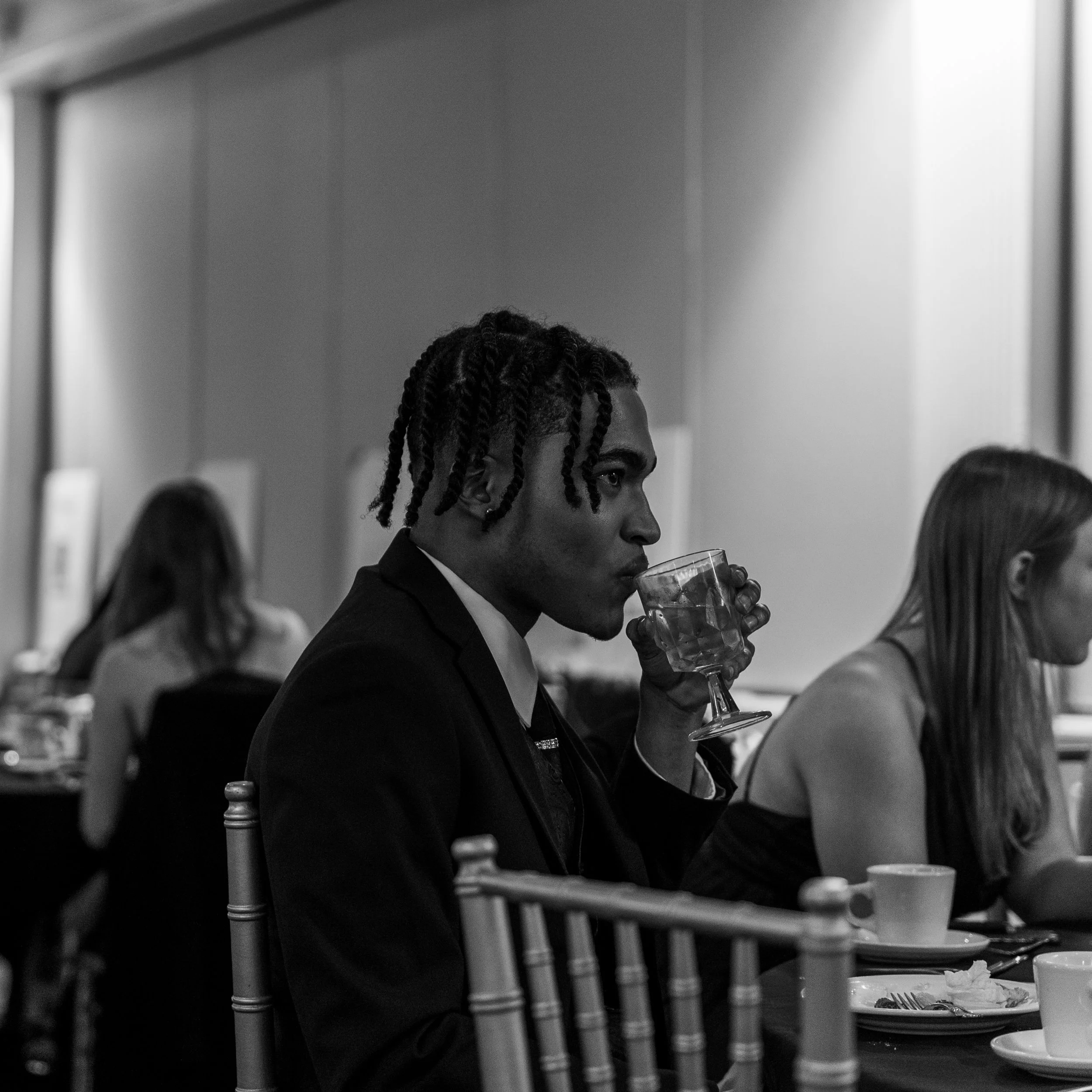 A man with styled braids dressed in a dark suit, sitting at a dining table, drinking from a glass of water, in a black-and-white indoor setting.