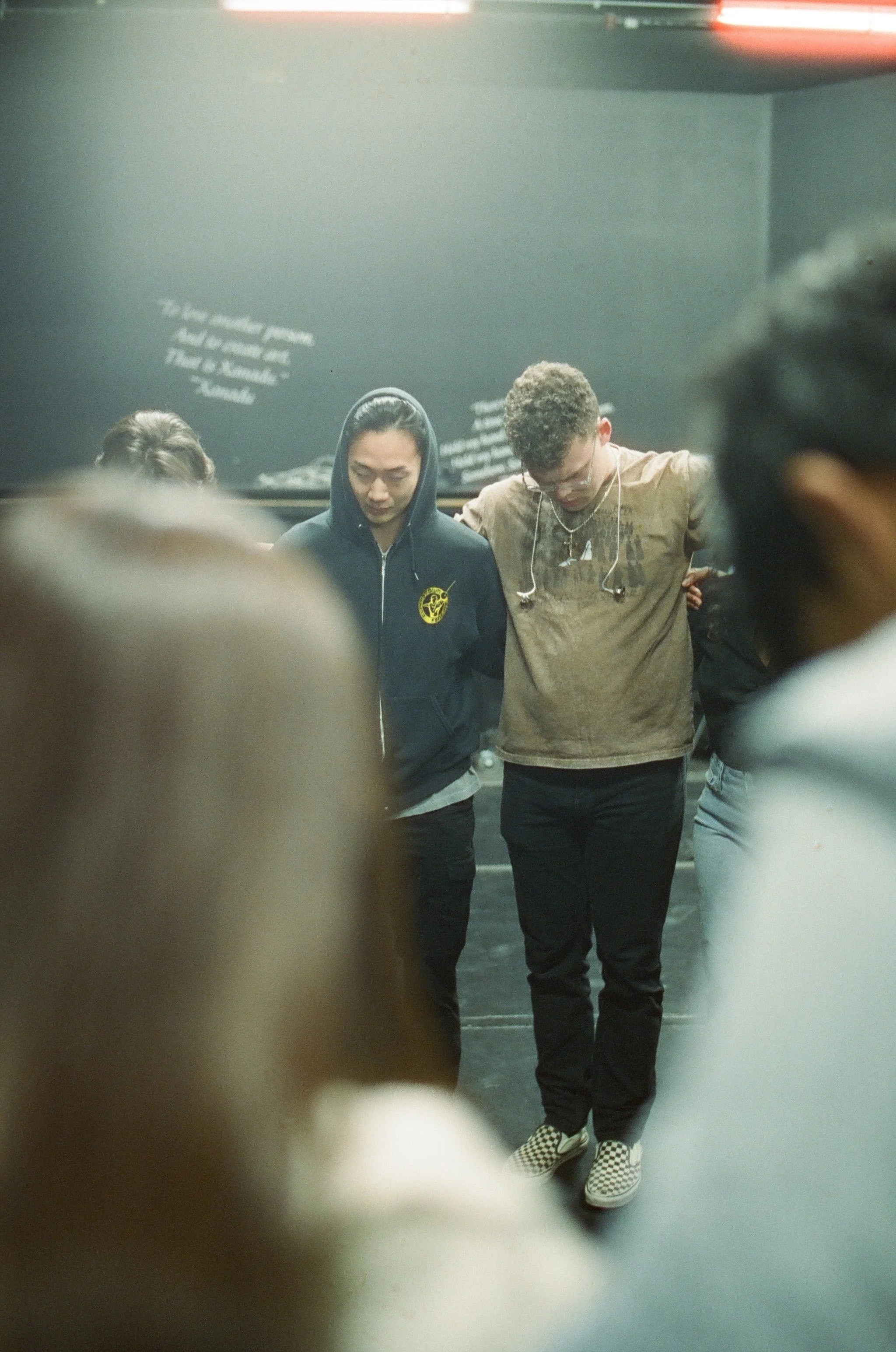 People standing in a circle with their heads bowed and eyes closed, possibly praying or meditating in a dimly lit room.