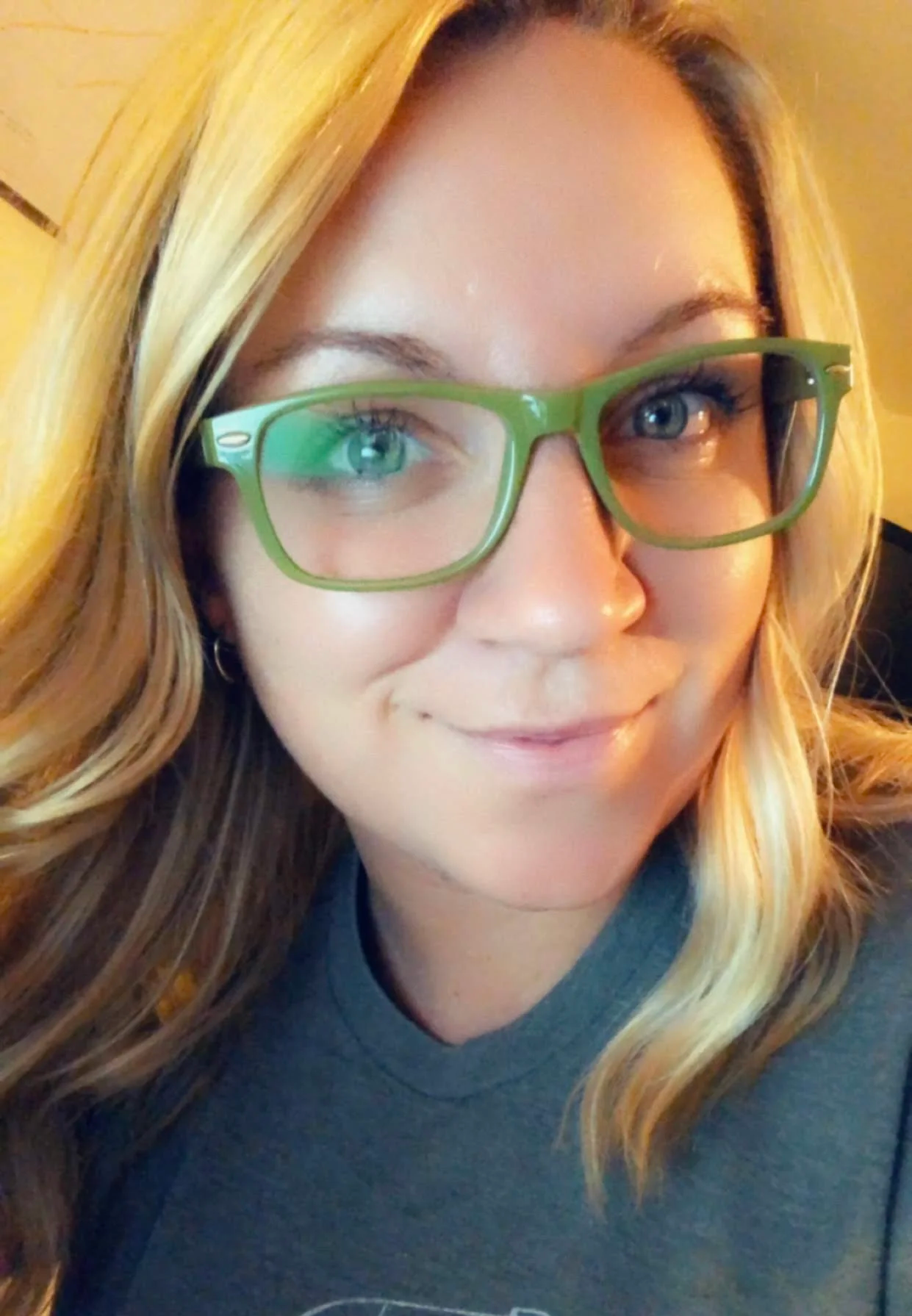 Close-up of a smiling woman with blonde hair wearing green glasses and a dark-colored shirt, looking at the camera.