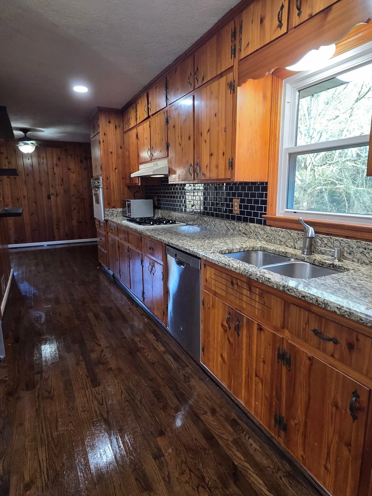 Wood-paneled kitchen with granite countertops, black tile backsplash, stainless steel dishwasher, oven, and a double sink, lit by a window and ceiling lights.