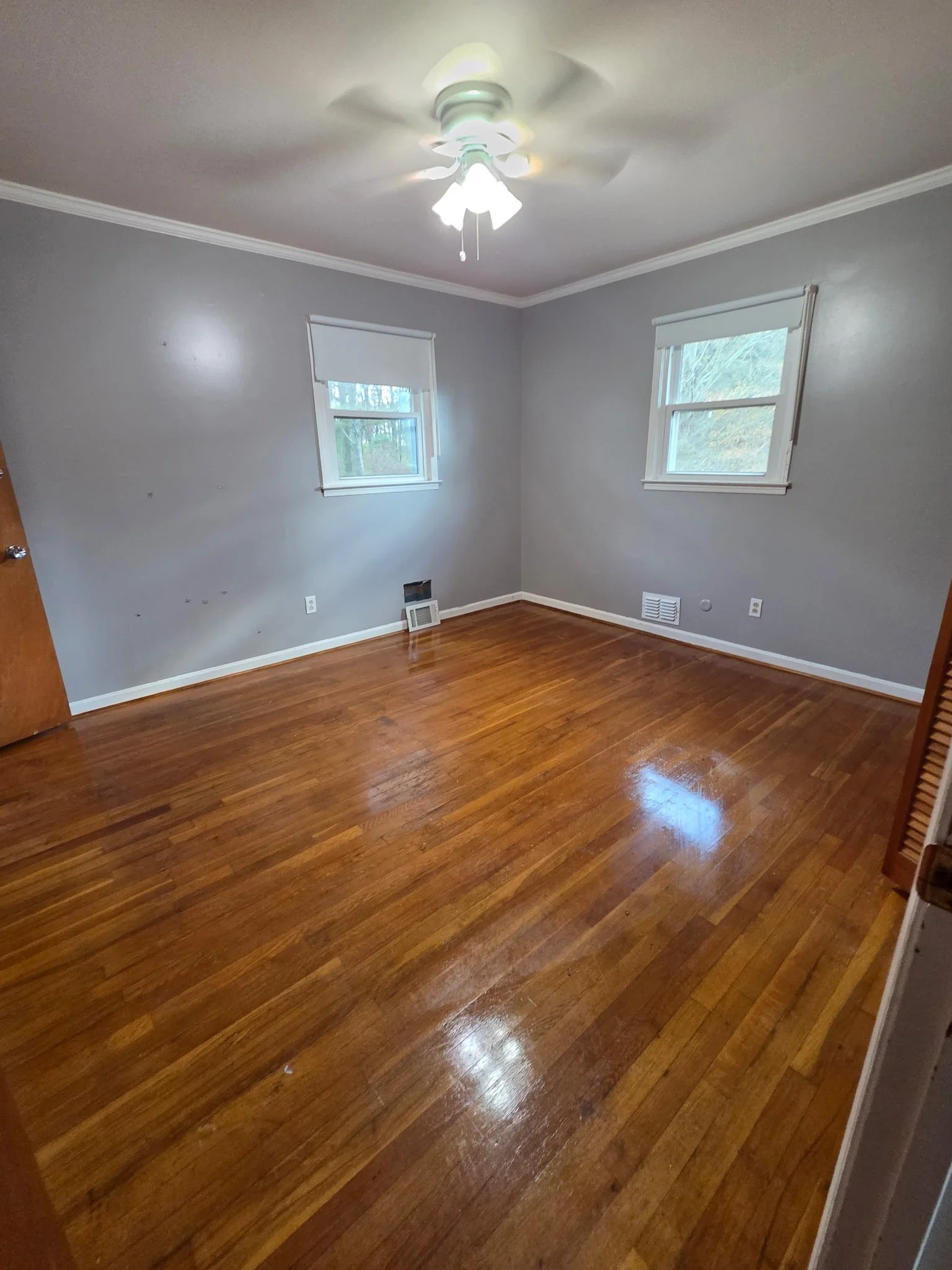Empty bedroom with hardwood floors, light gray walls, two windows with white blinds, a ceiling fan with light, and a closed wooden door.