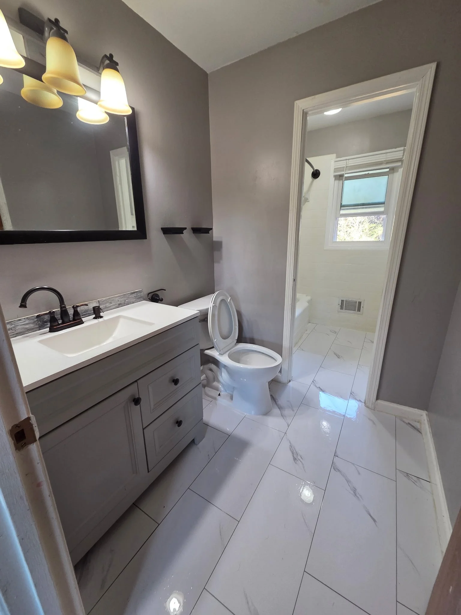 Modern bathroom with gray walls, white marble floor tiles, a white vanity with a black faucet, a large mirror with a black frame, and a toilet. In the background, a shower area with a window is visible.