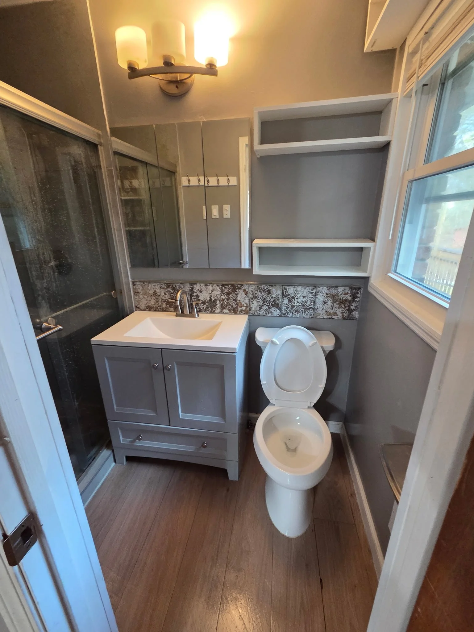 A small bathroom with a vanity, toilet, and shower stall, gray walls, and a window with blinds.