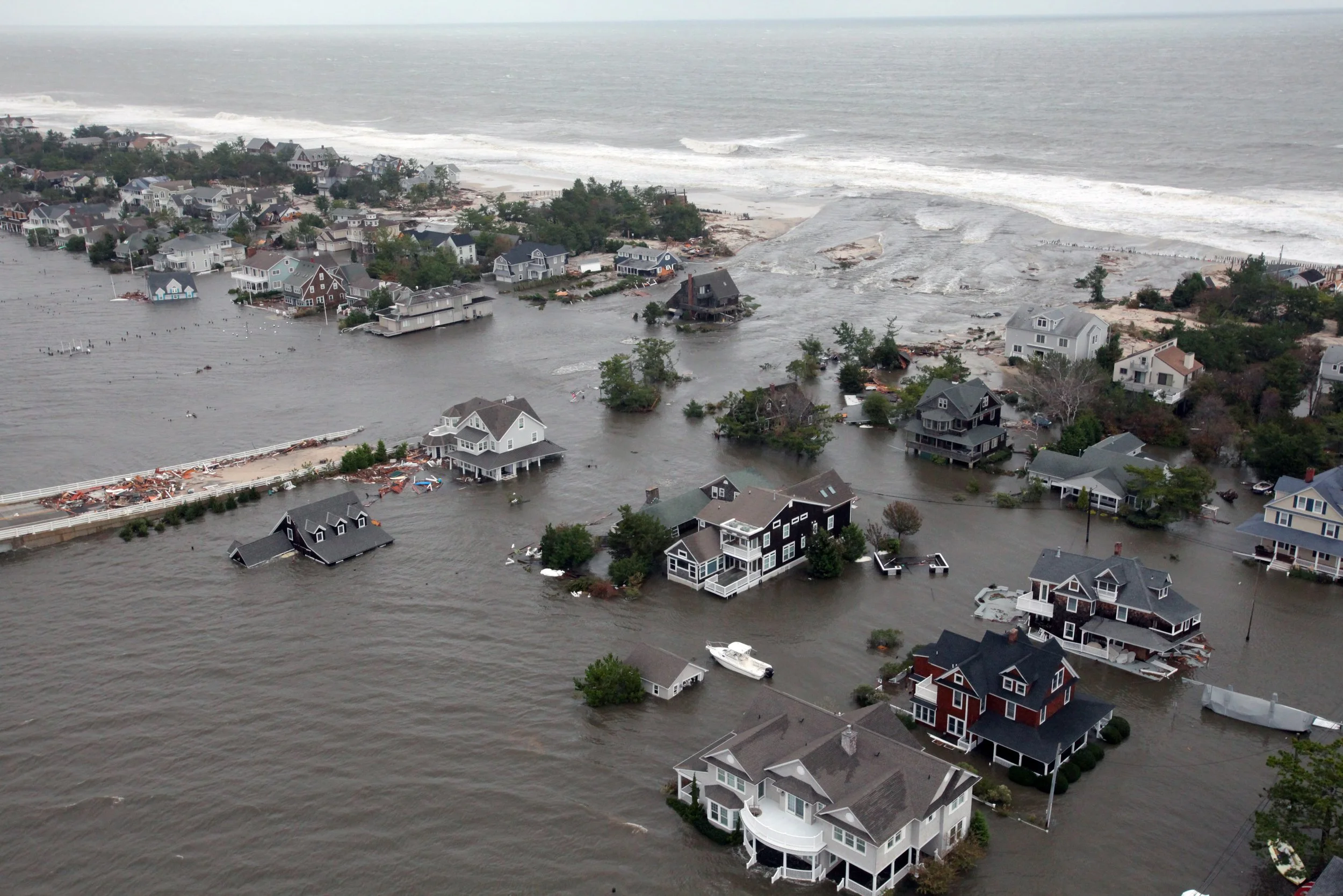 An aerial view of a coastal neighborhood submerged in floodwaters after a storm, with houses partially underwater and debris scattered around.