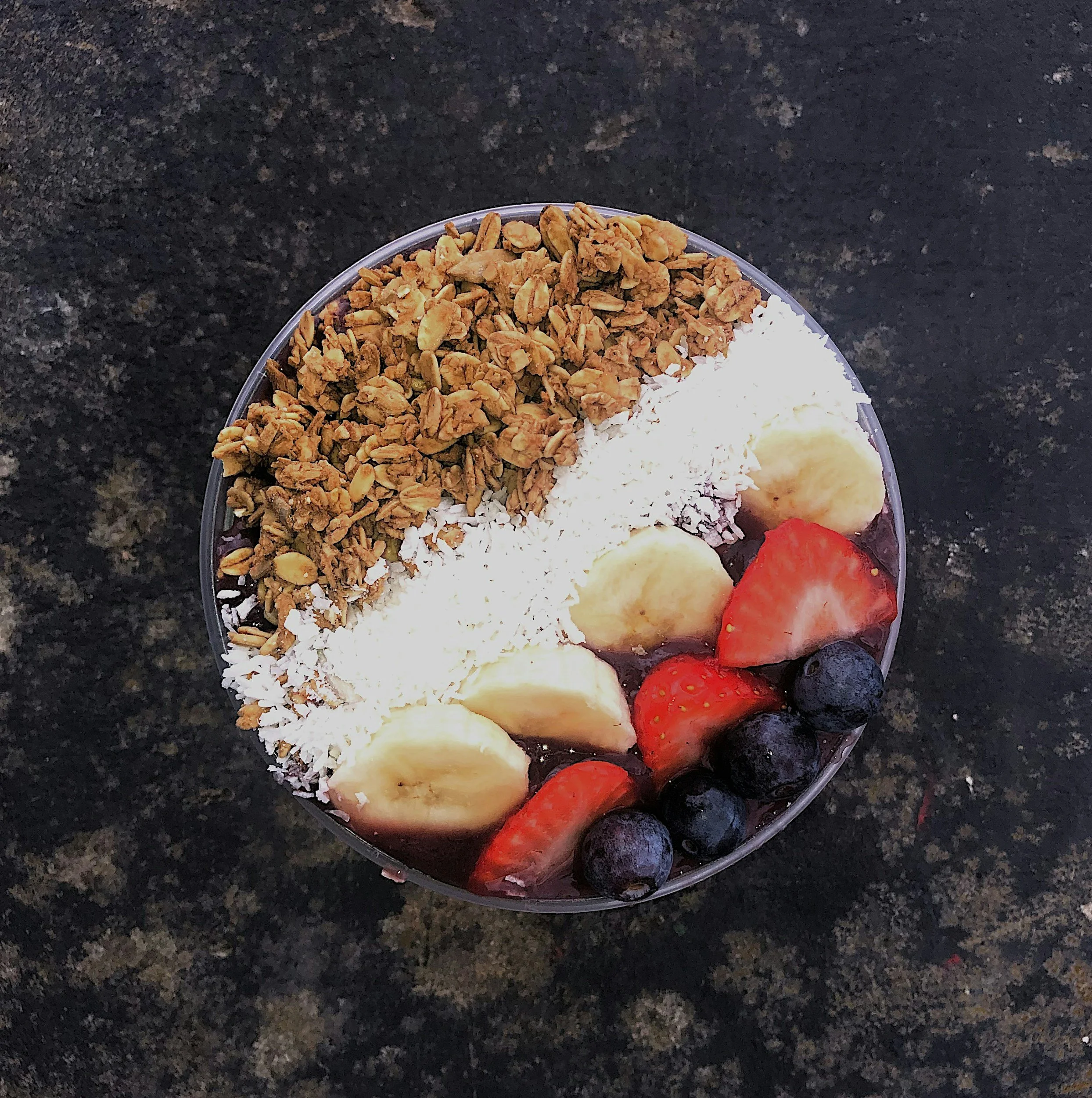 A bowl of acai smoothie topped with granola, banana slices, shredded coconut, strawberry slices, and blueberries, placed on a dark, textured surface.
