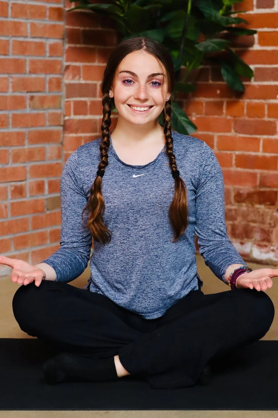 A young woman with brown hair in two braids is sitting cross-legged on a black yoga mat indoors, smiling with her hands on her knees in a meditation pose. She is wearing a blue long-sleeve athletic shirt and black pants, with a background of a brick wall and green plants.